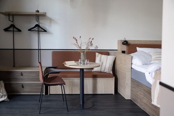 A compact hotel room with a bed, a small round table with a vase of flowers, a sofa, a chair, and a hanging shelf with clothes.