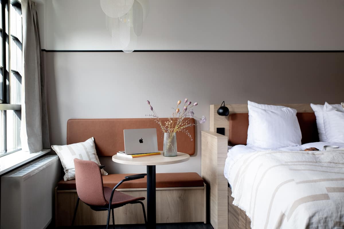 Hotelkamer met een bed, een bureau met een laptop, een stoel, een kleine tafel met een vaas en een raam met gordijnen.