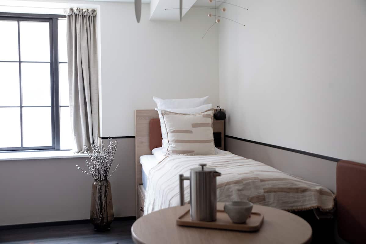 Een keurig opgeruimde hotelkamer met een enkel bed, een nachtkastje met waterkoker en beker, een vaas met bloemen en uitzicht op het buitenleven door een raam met gordijnen.