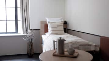 A neatly arranged hotel room with a single bed, a bedside table with a kettle and cup, a vase with flowers, and a view of the outdoors through a window with curtains.