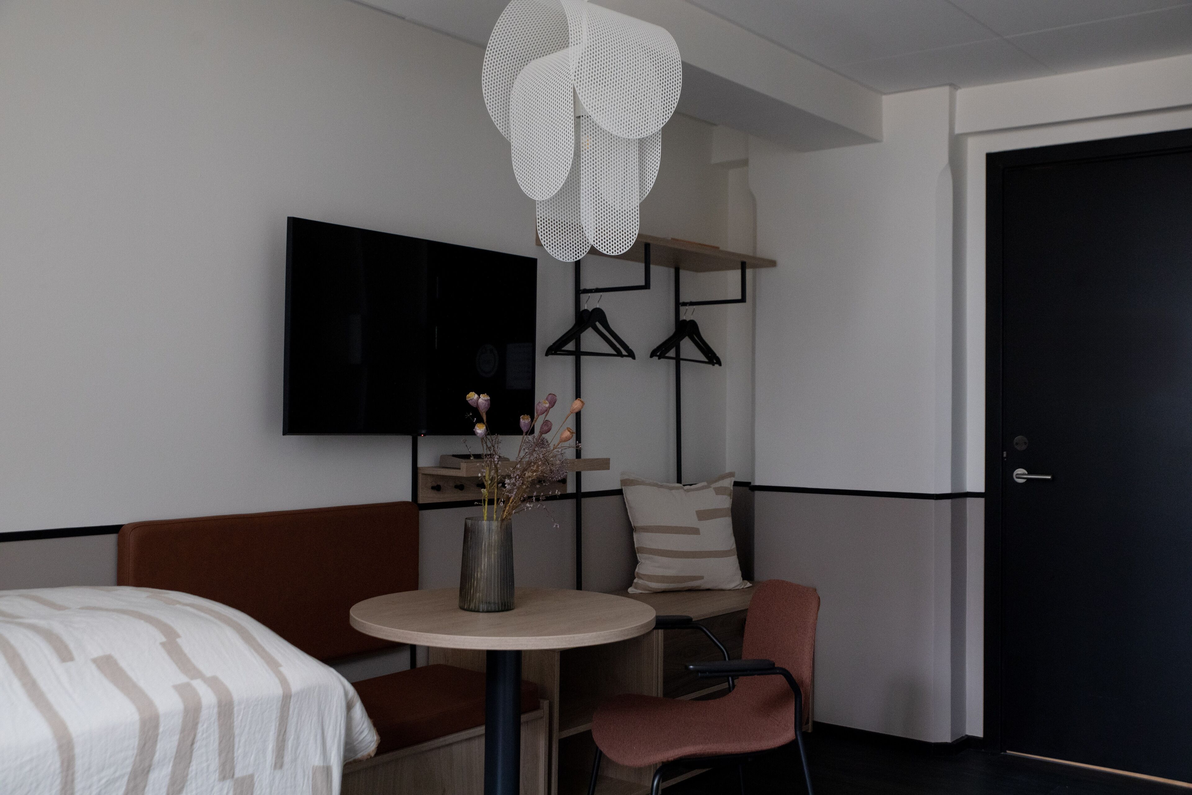 A hotel room with a bed, a round table with a vase, a chair, a TV mounted on the wall, and a door.