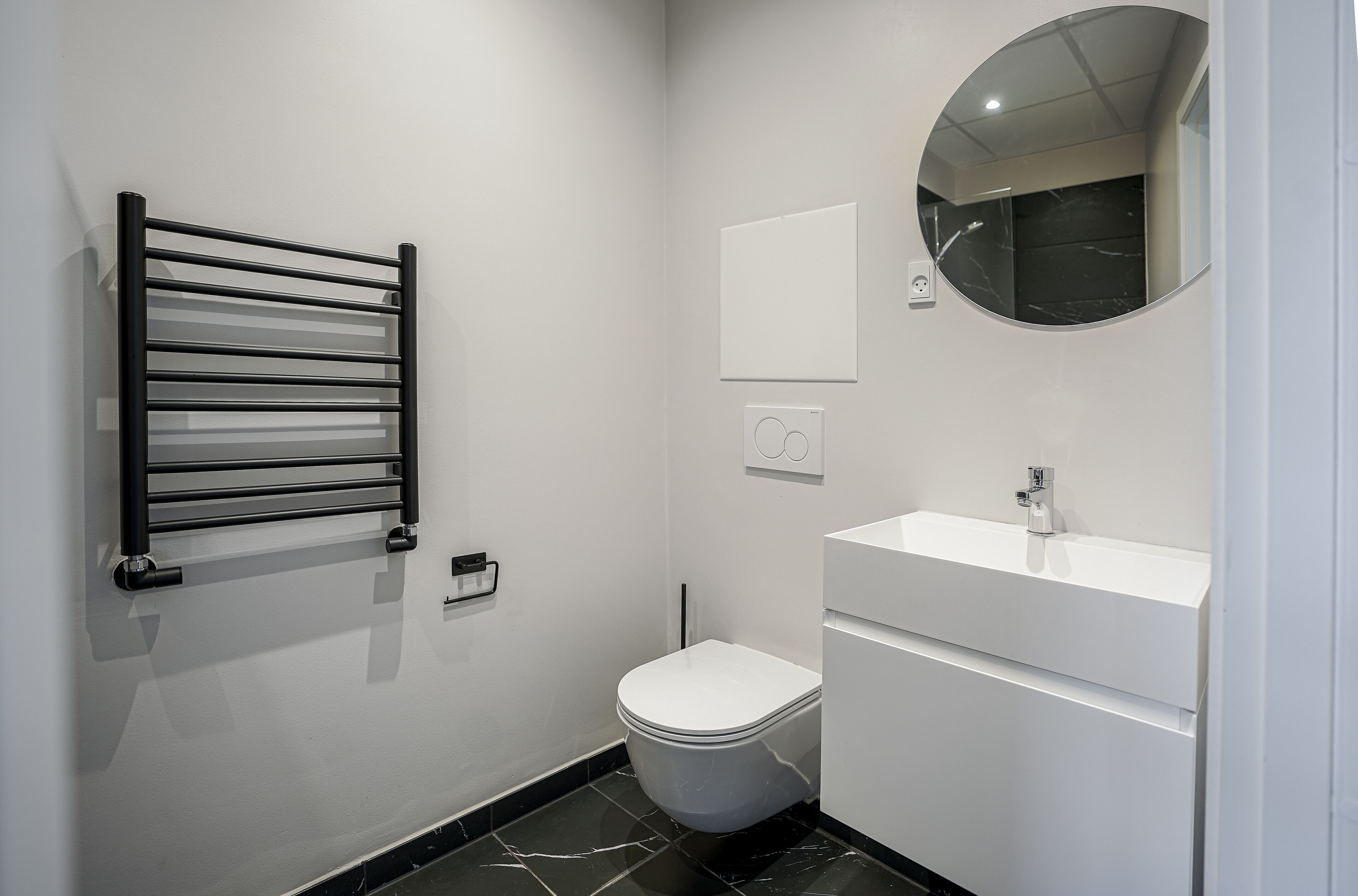 A modern bathroom with a black towel rack, a white toilet, a white sink, and a round mirror.