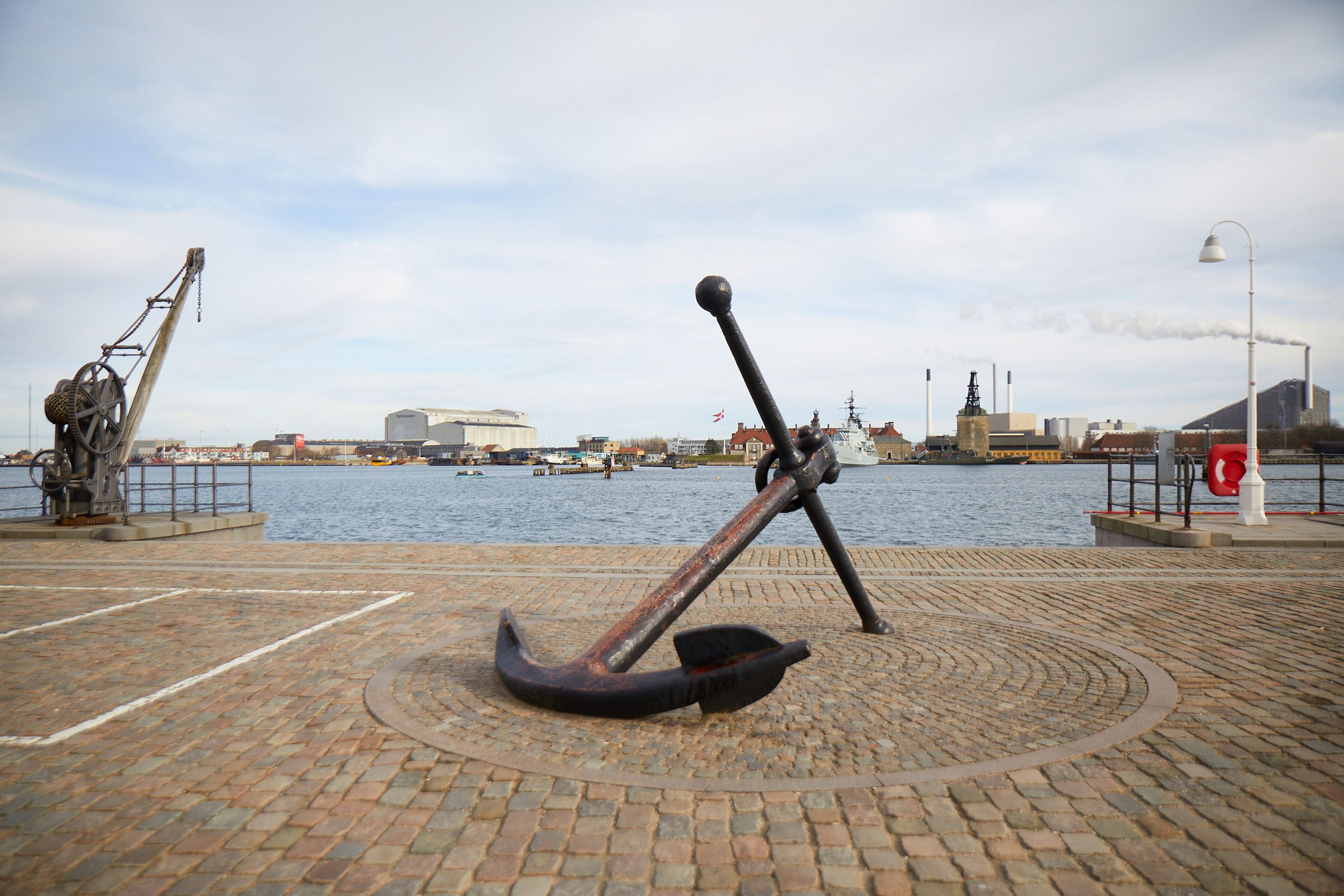 A waterfront area with a large anchor sculpture in the foreground, a brick pathway, and a view of the water with boats and industrial buildings in the background.