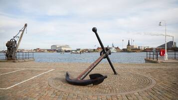 A waterfront area with a large anchor sculpture in the foreground, a brick pathway, and a view of the water with boats and industrial buildings in the background.