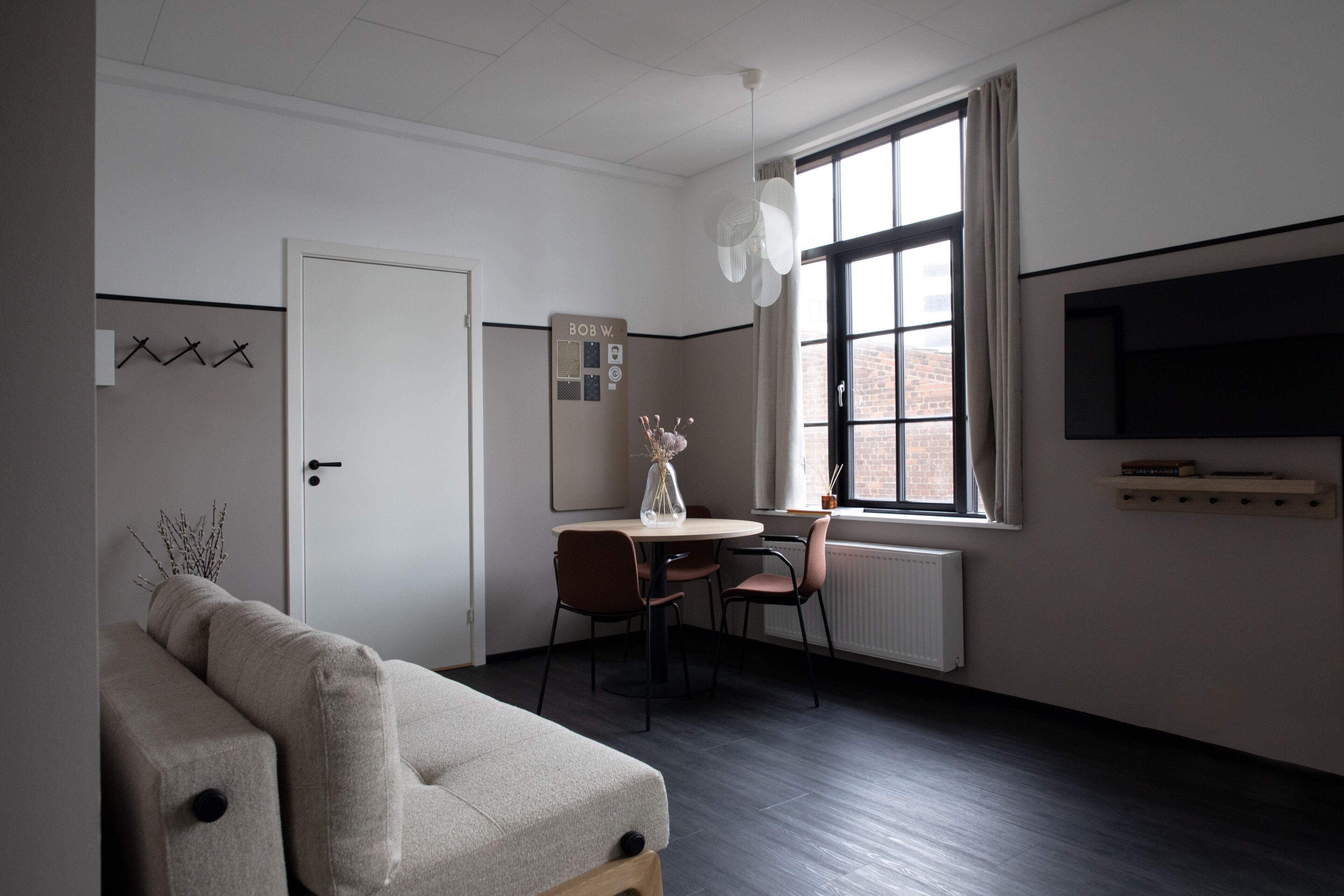 A modern, minimalist room with a sofa, a dining table with chairs, a large window with a view of a brick building, and a mounted television.