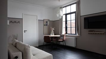 A modern, minimalist room with a sofa, a dining table with chairs, a large window with a view of a brick building, and a mounted television.