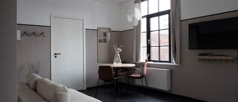Ett modernt, minimalistiskt rum med en soffa, ett matbord med stolar, ett stort fönster med utsikt över en byggnad med tegelstensfasad och en väggmonterad tv.