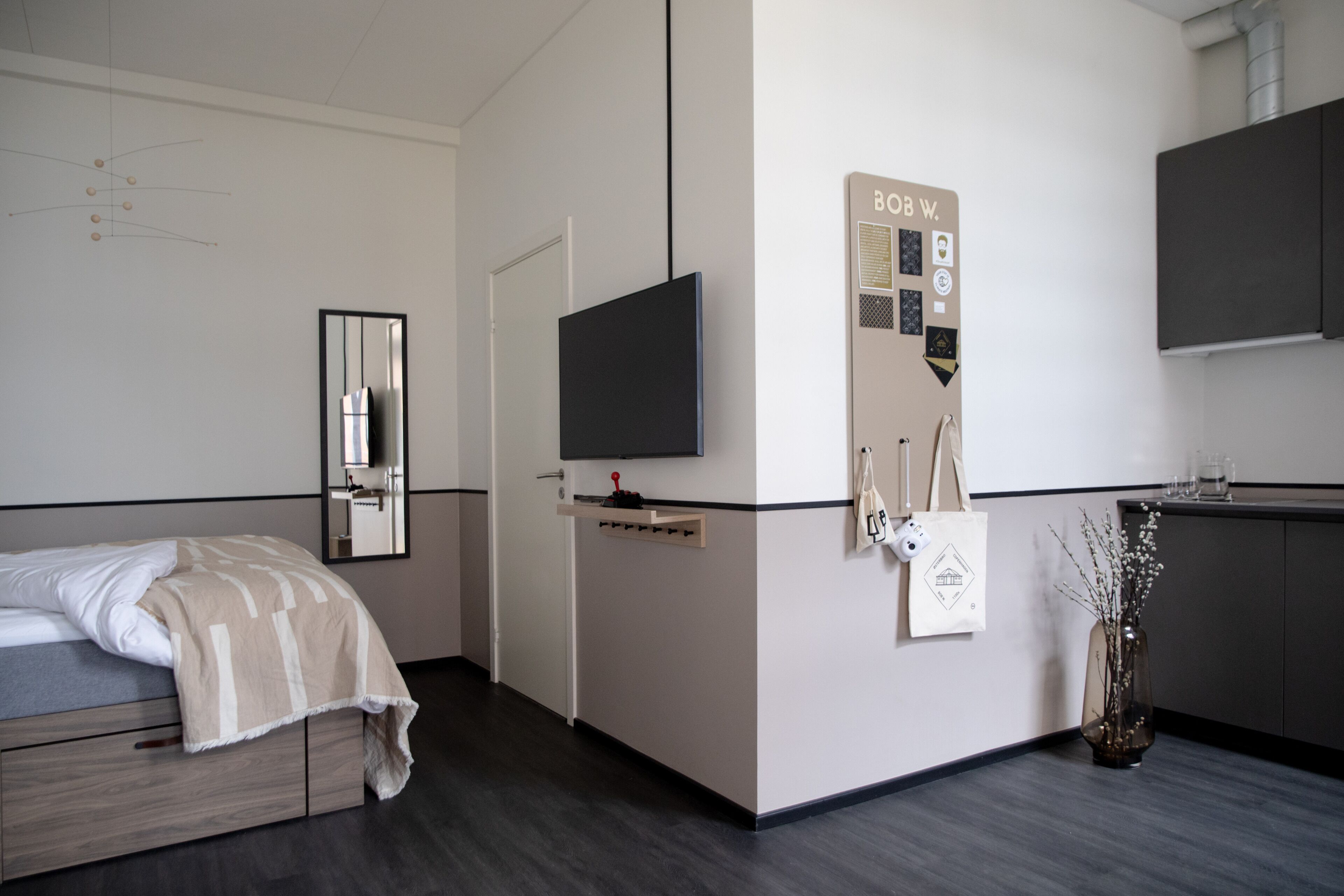 A modern hotel room with a bed, a TV, a small kitchenette, and a mirror.