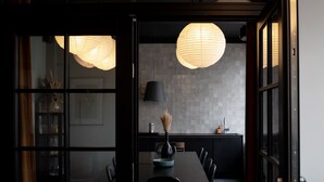Un comedor moderno con una mesa de madera oscura, sillas negras y un jarrón de flores secas. Un farolillo blanco grande y redondo cuelga del techo.
