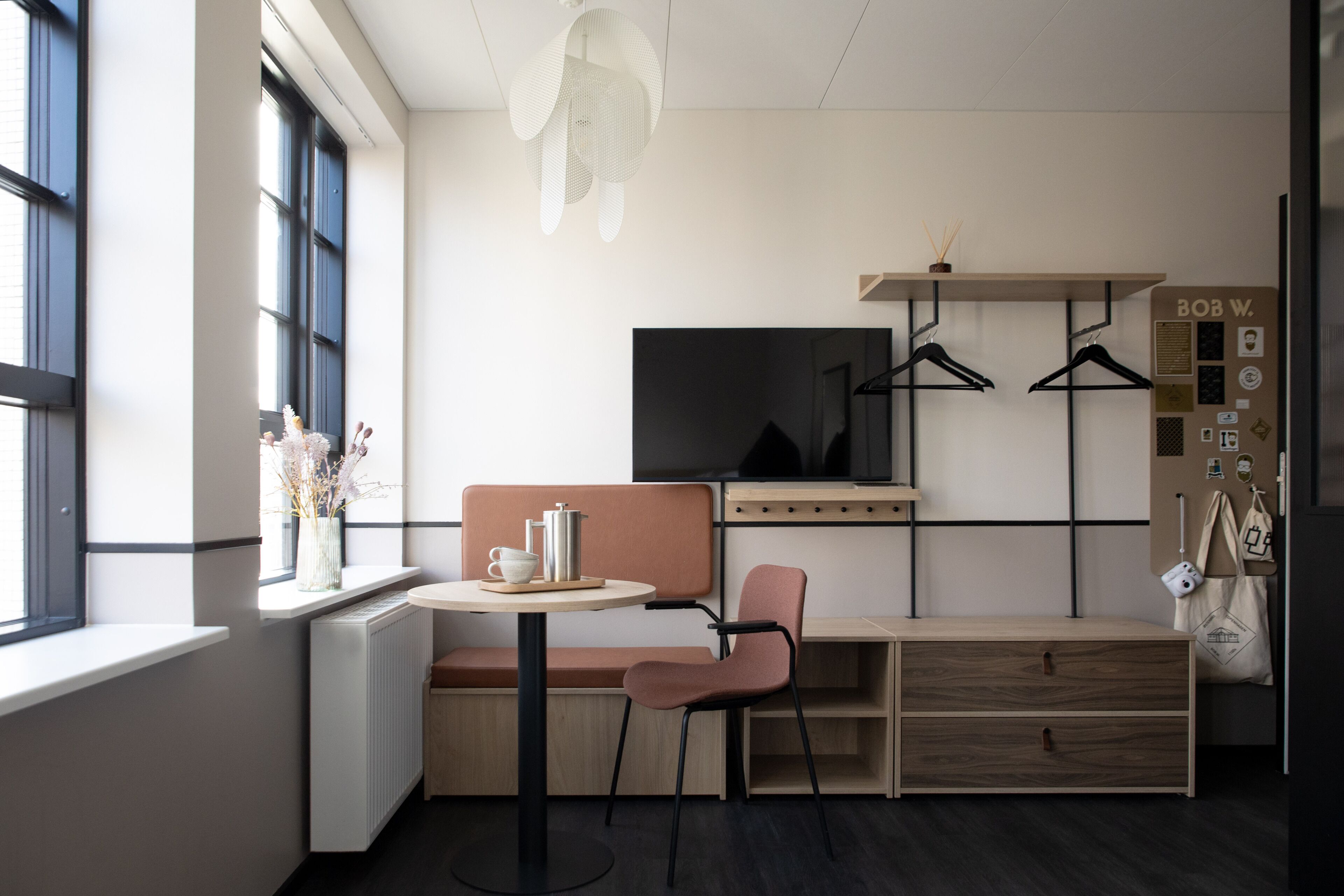 A modern, well-lit room with a large window, a round table with a chair, a bench, a television, and a wardrobe with a hanging organizer.