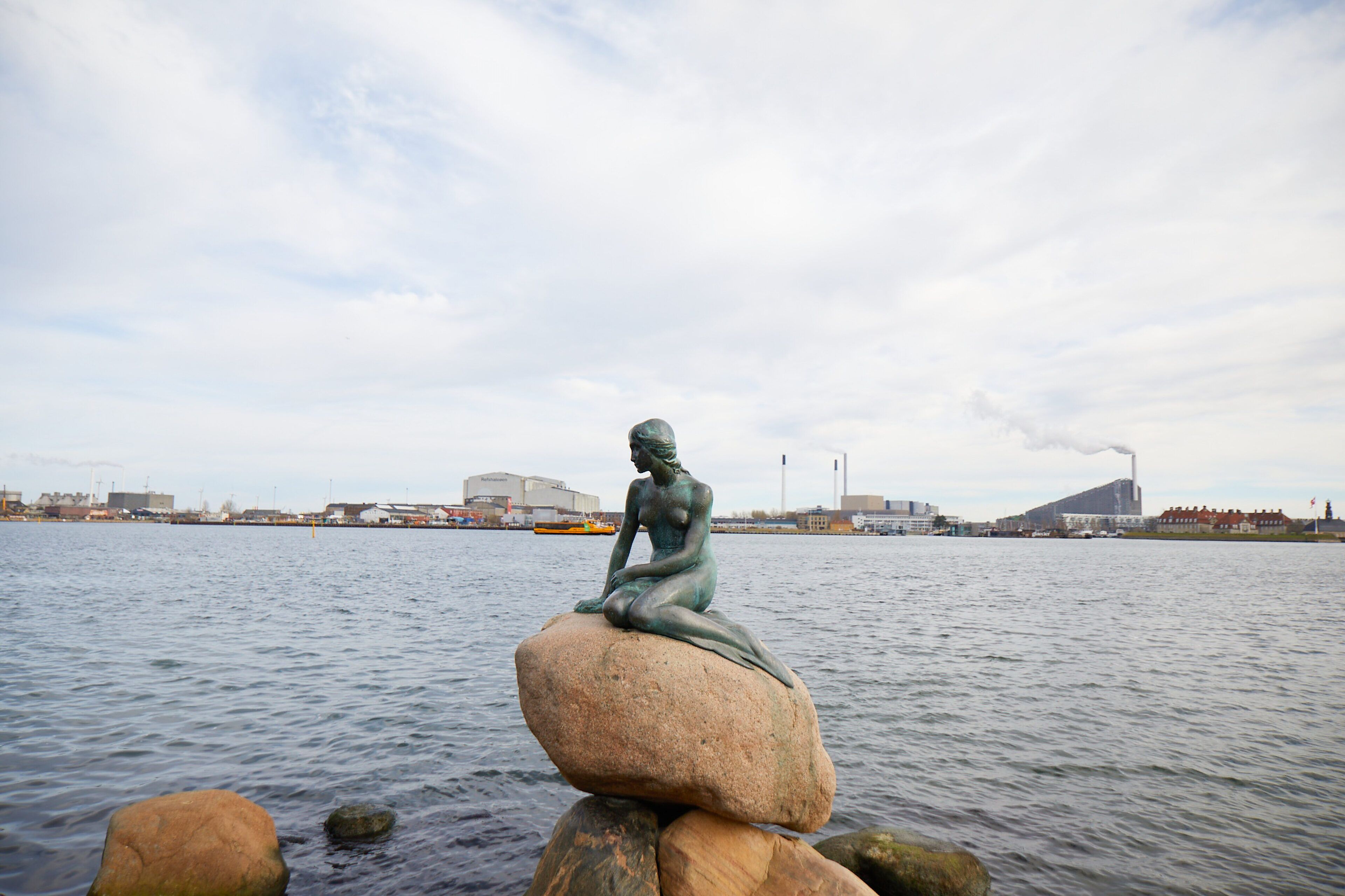 Een standbeeld van een zittend figuur op een rots aan een waterlichaam, met industriële gebouwen op de achtergrond.