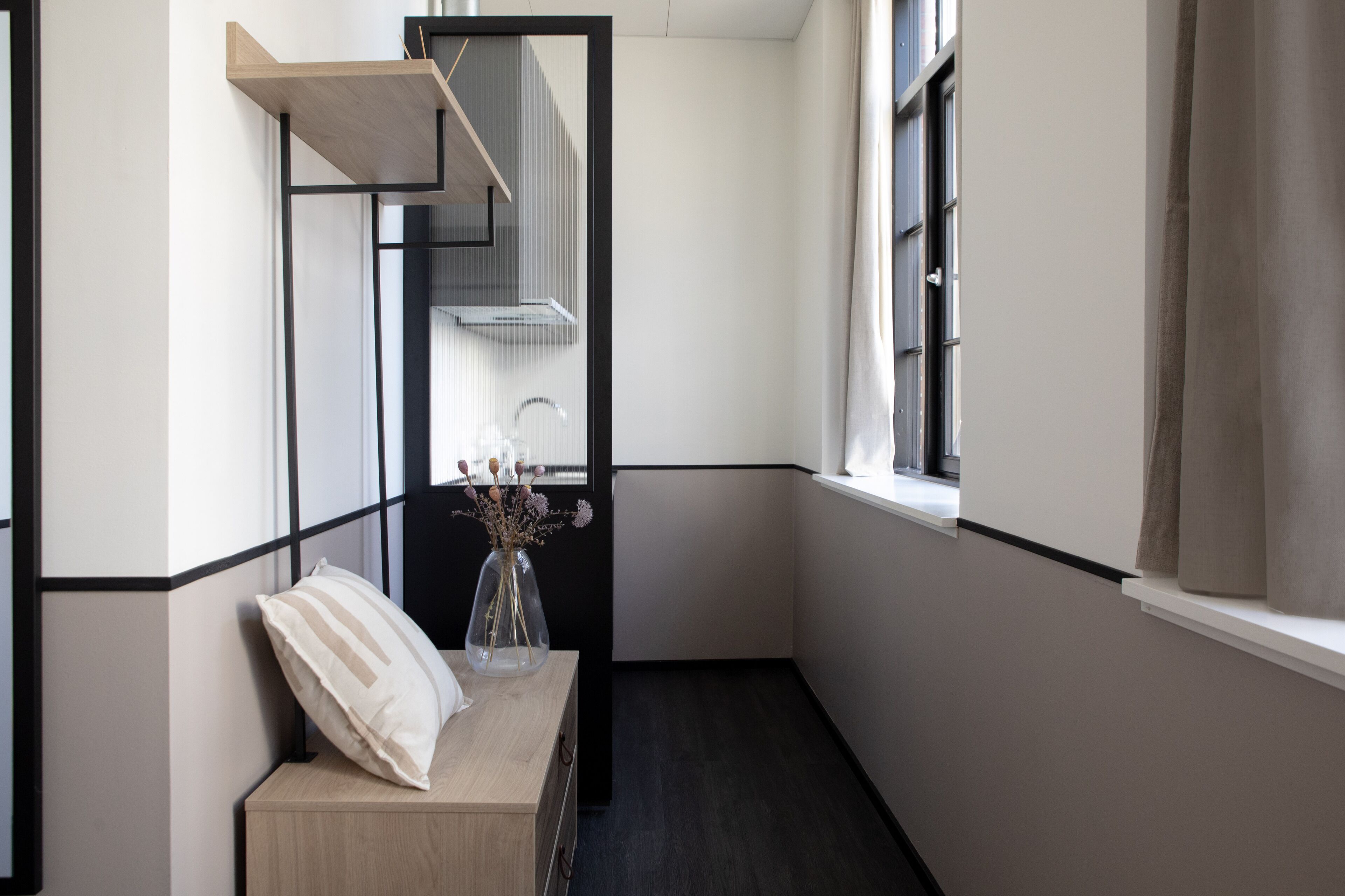 A modern hotel room with a minimalist design, featuring a wooden bench, a vase with flowers, and a large window providing natural light.