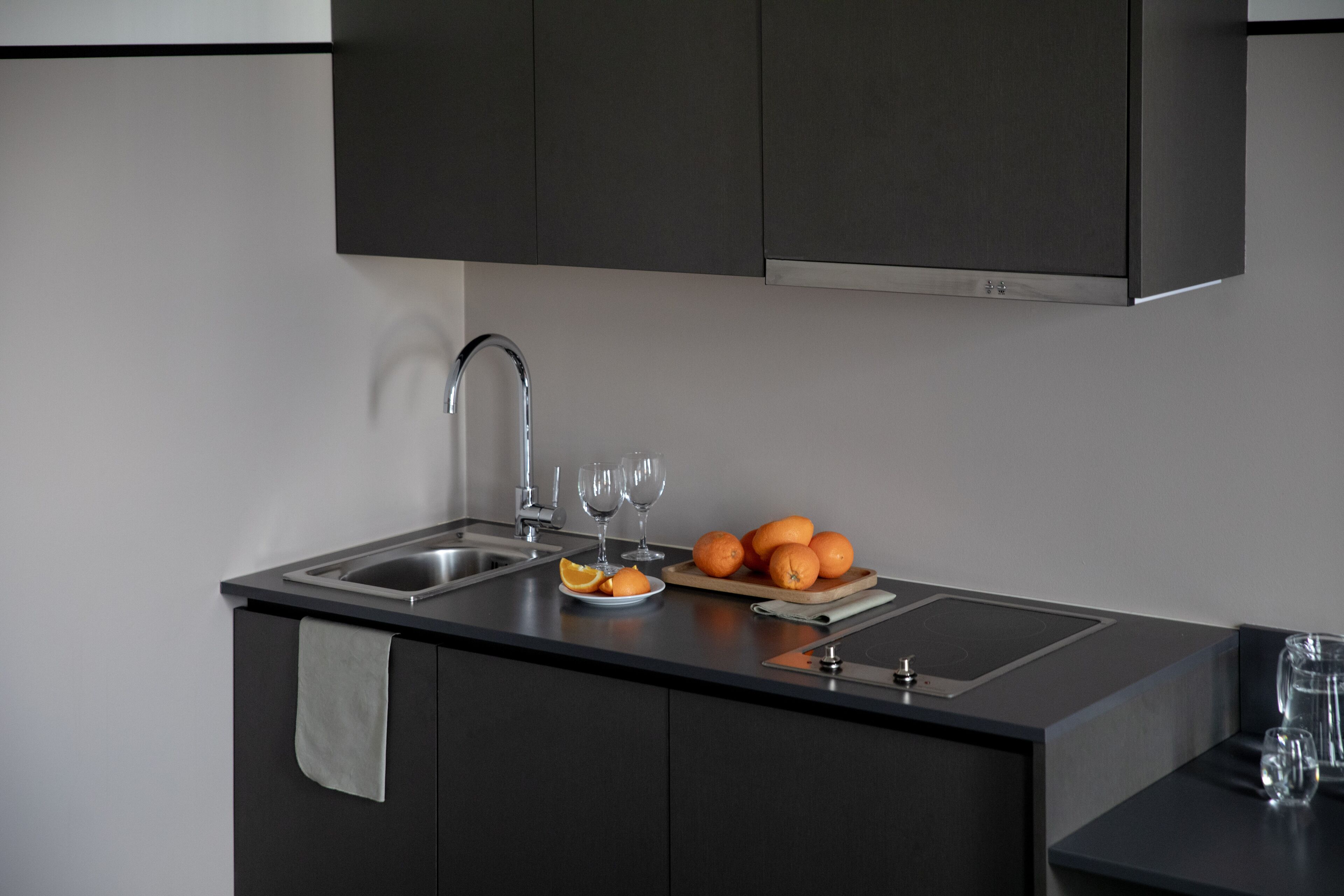 Une cuisine moderne dotée d’un évier en acier inoxydable, de meubles de couleur sombre et d’un plan de travail agrémenté d’une planche à découper, d’oranges et de verres à vin.