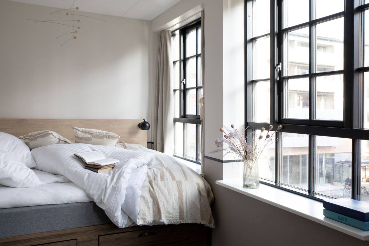 Een slaapkamer met een bed, een raam met uitzicht op gebouwen en een nachtkastje met een vaas.
