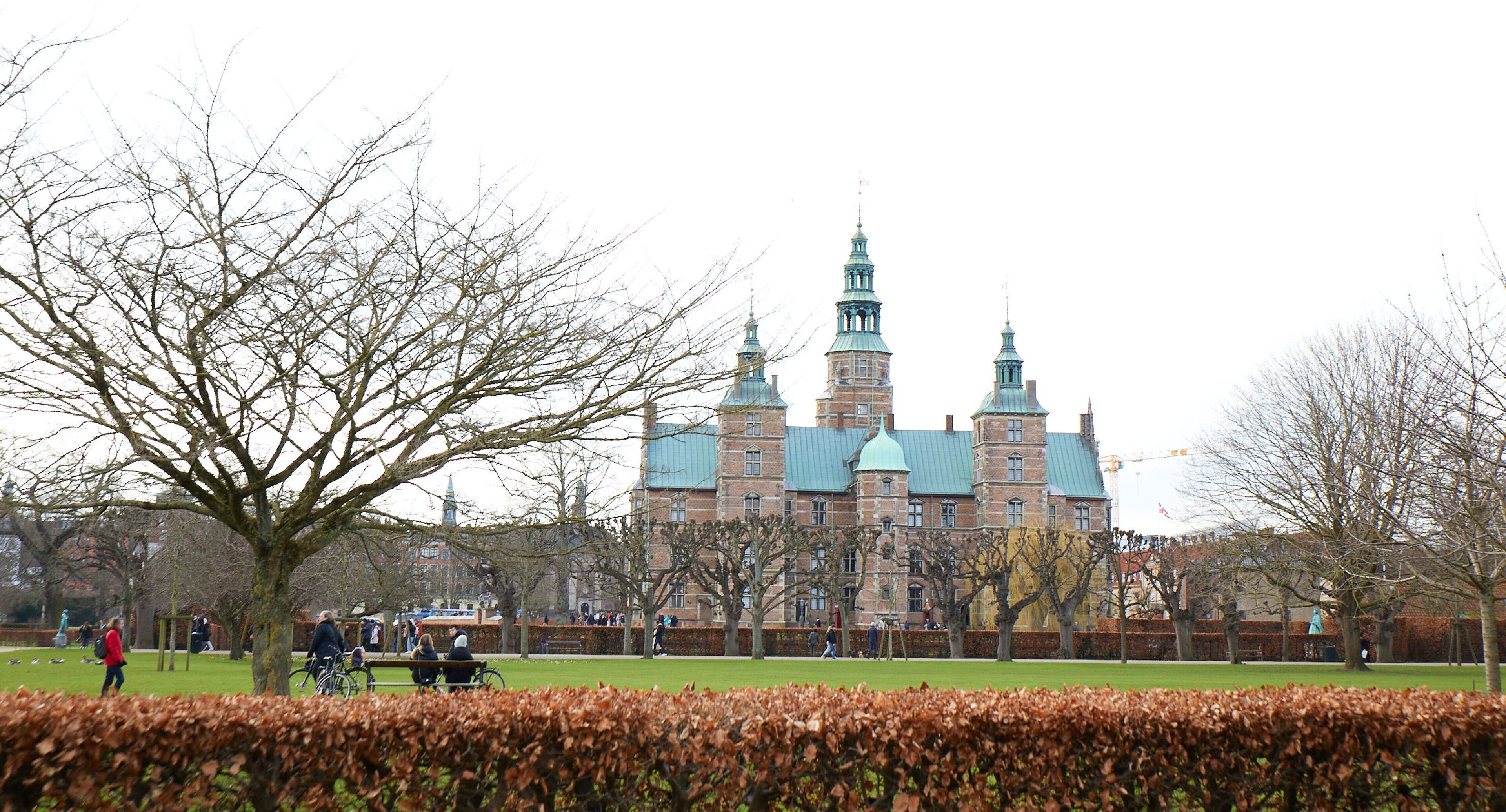 A large historic building with multiple towers and a green roof, surrounded by a park with benches and people.