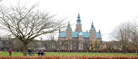 A large historic building with multiple towers and a green roof, surrounded by a park with benches and people.