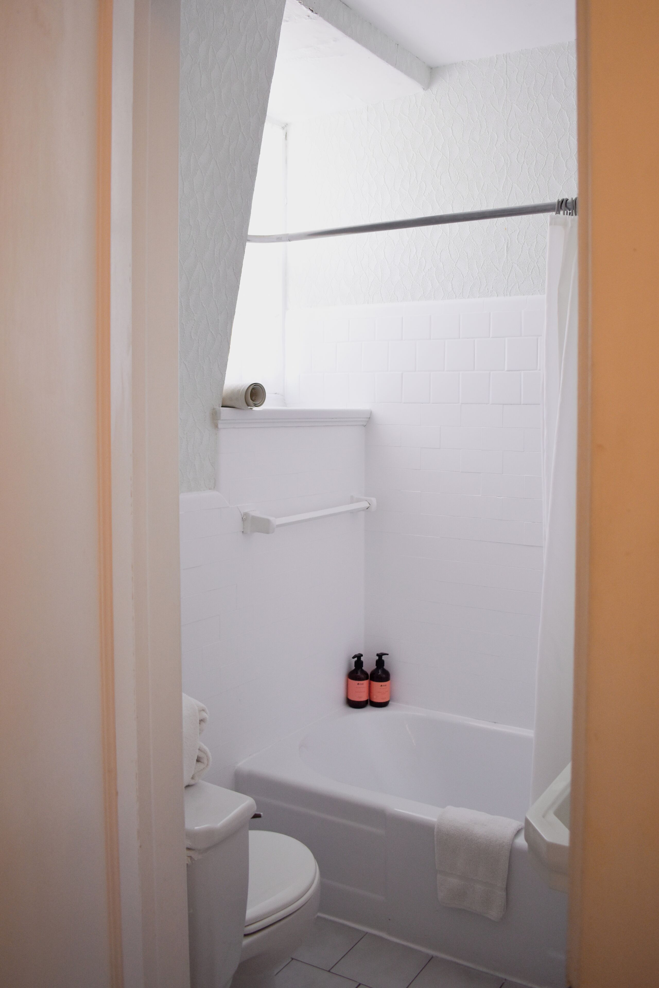 Une petite salle de bain blanche, équipée de toilettes, d’une baignoire et d’un espace douche. Deux bouteilles sont posées sur le mur.