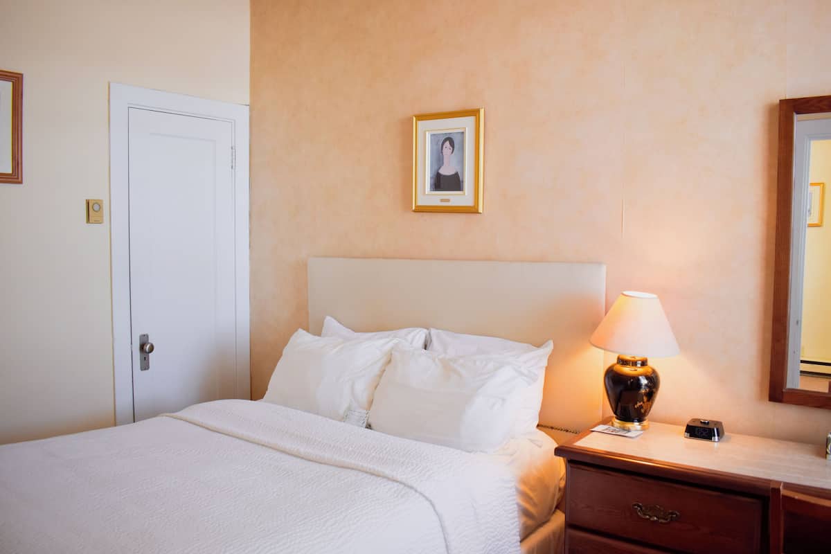 A neatly made bed with white linens, a bedside lamp, a framed picture on the wall, and a wooden nightstand with a drawer.