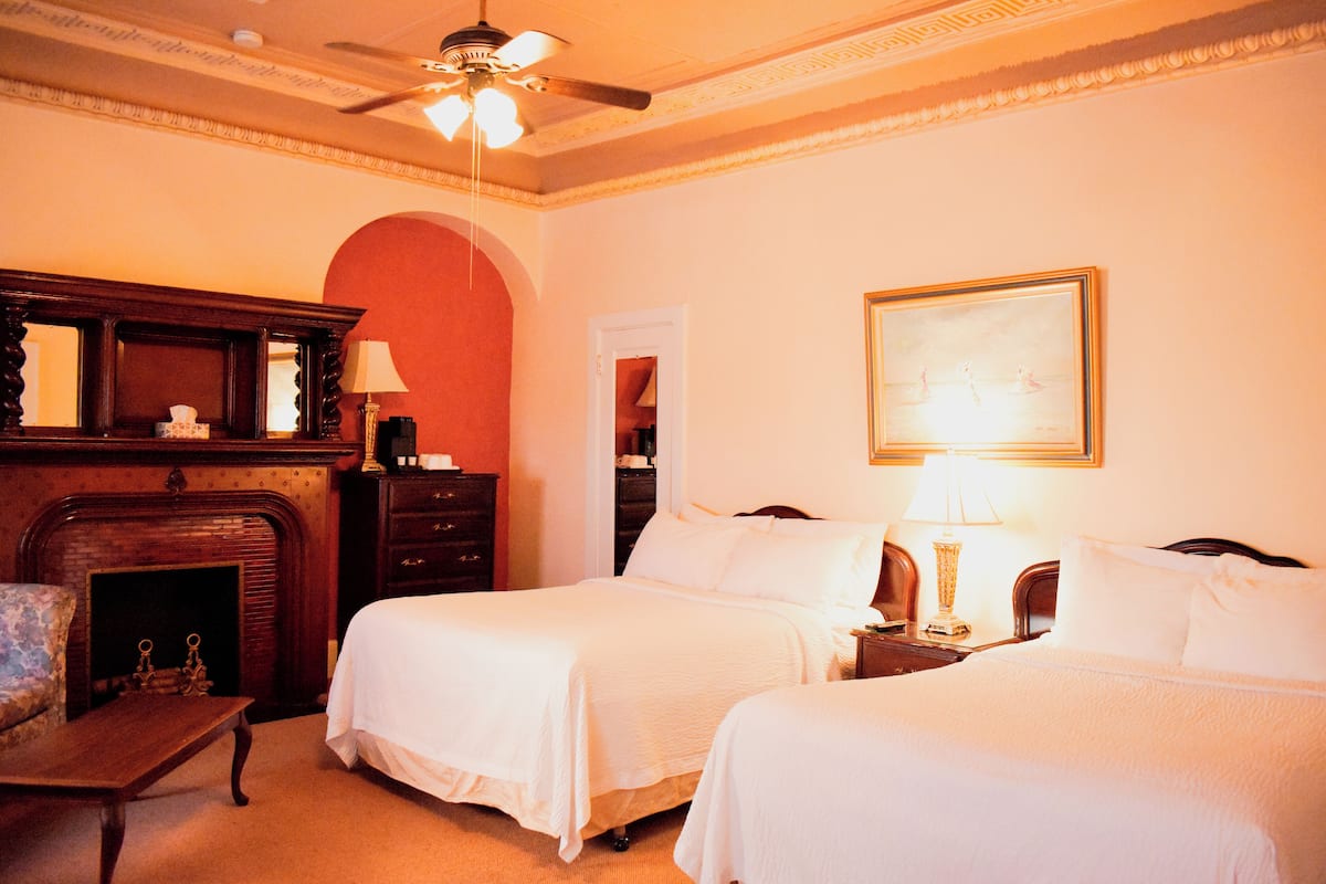 A hotel room with two beds, a fireplace, a ceiling fan, and a painting on the wall.