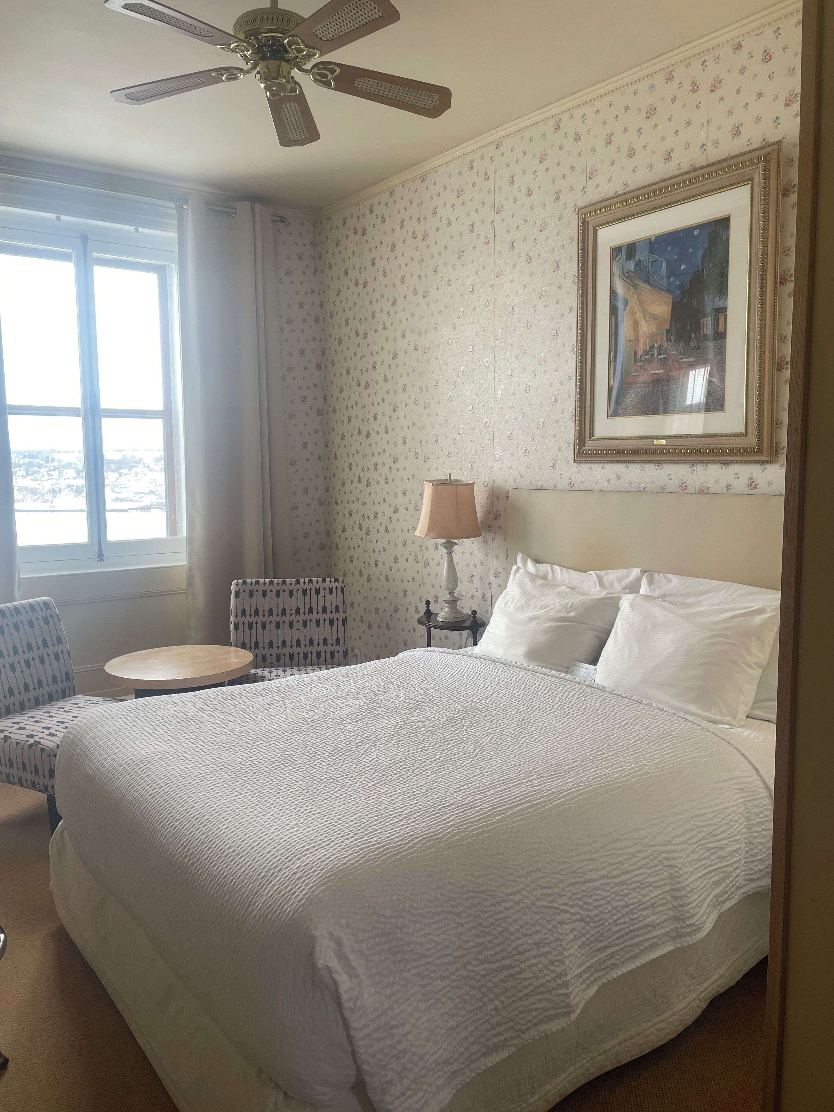 Habitación de hotel con una cama grande, un ventilador de techo, un cuadro enmarcado, una lámpara y vistas al exterior a través de una ventana.