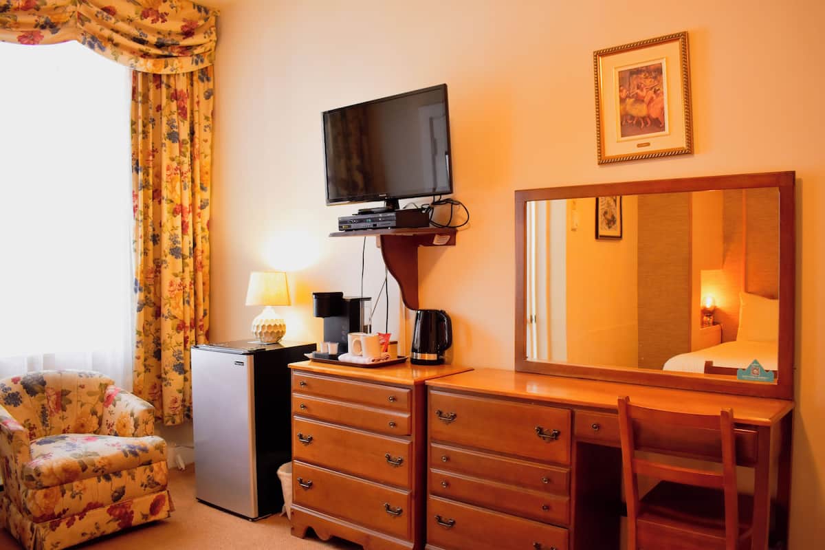A hotel room with a television mounted on the wall, a dresser with drawers, a small refrigerator, a floral armchair, and a bedside table with a lamp and a cup.
