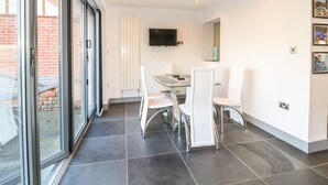 Ein stilvoller Essbereich mit Glastisch und weißen Stühlen, einem an der Wand befestigten Flachbildfernseher und Blick durch große Glasüren.