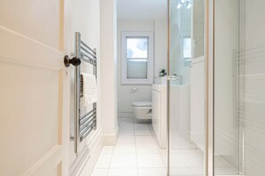 Un couloir étroit mène à une salle de bain équipée de toilettes, d’un lavabo et d’une douche à parois de verre.