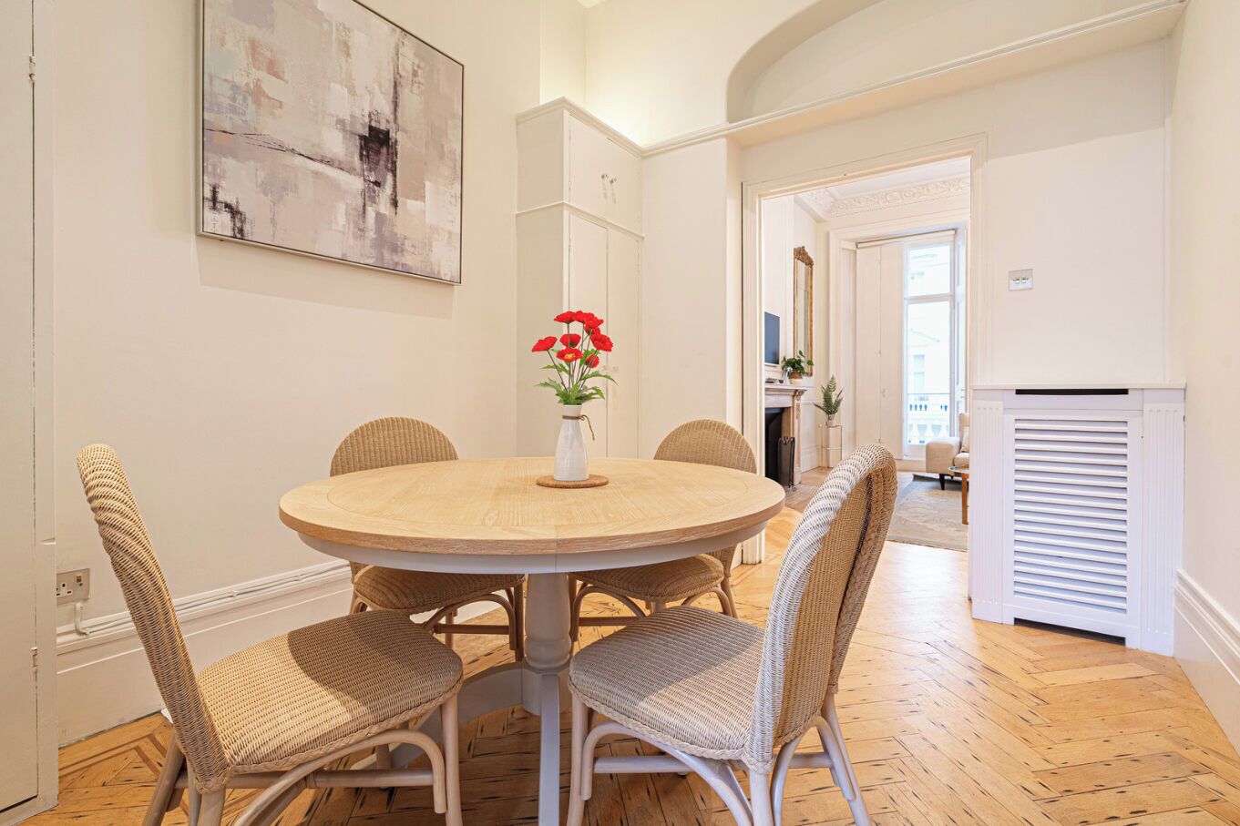 A dining area with a round table, wicker chairs, and a vase with red flowers.