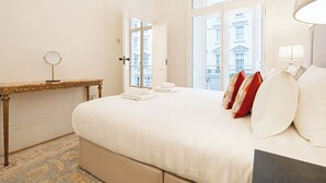 A hotel room with a large bed, a bedside table with a mirror, and a view of buildings through the windows. - Beautiful Belgravia by Underthedoormat (London)