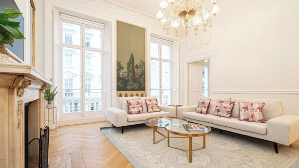 A spacious living room with a fireplace, two sofas with patterned cushions, a glass coffee table, and a large window offering a view of buildings. - Beautiful Belgravia by Underthedoormat (London)