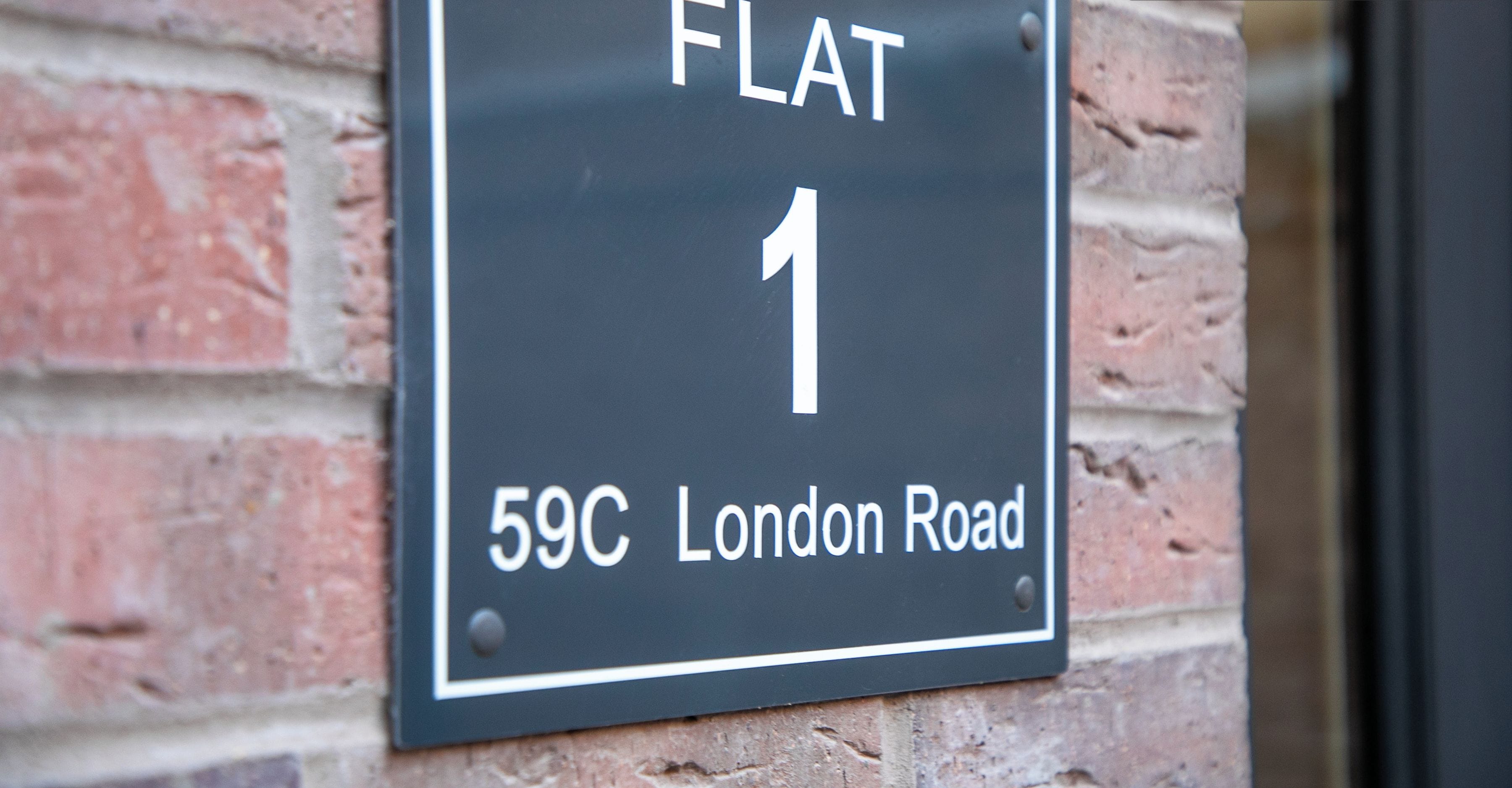A black sign with white lettering indicating "FLAT 1" and "59C London Road" on a brick wall.