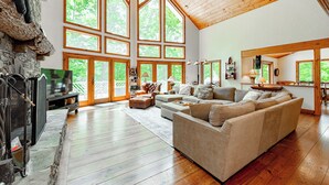 A spacious living room with a large wooden ceiling, a stone fireplace, and a large window overlooking greenery.