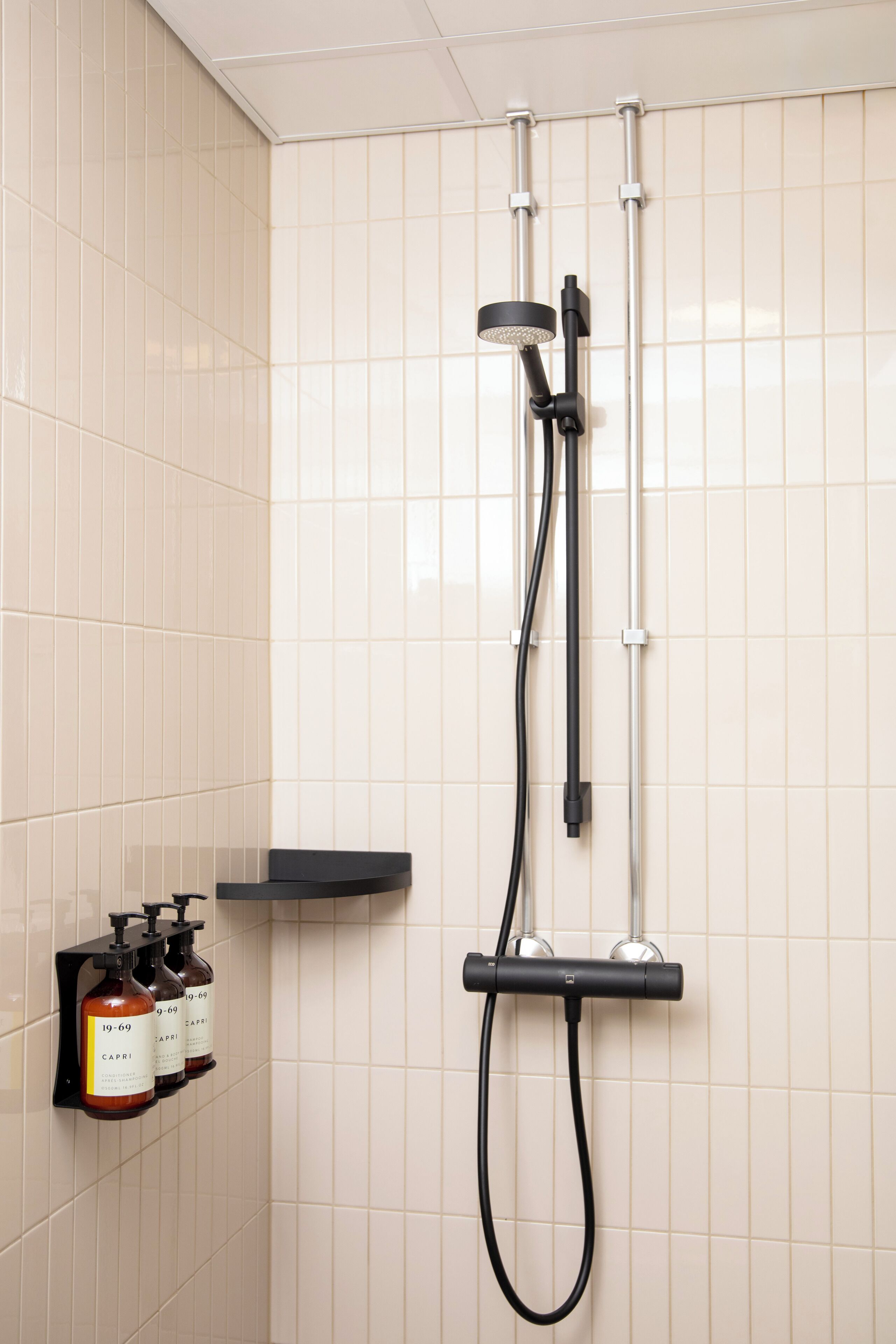 A shower with a handheld showerhead and a handheld hose. There are three bottles of shampoo on a black shelf.