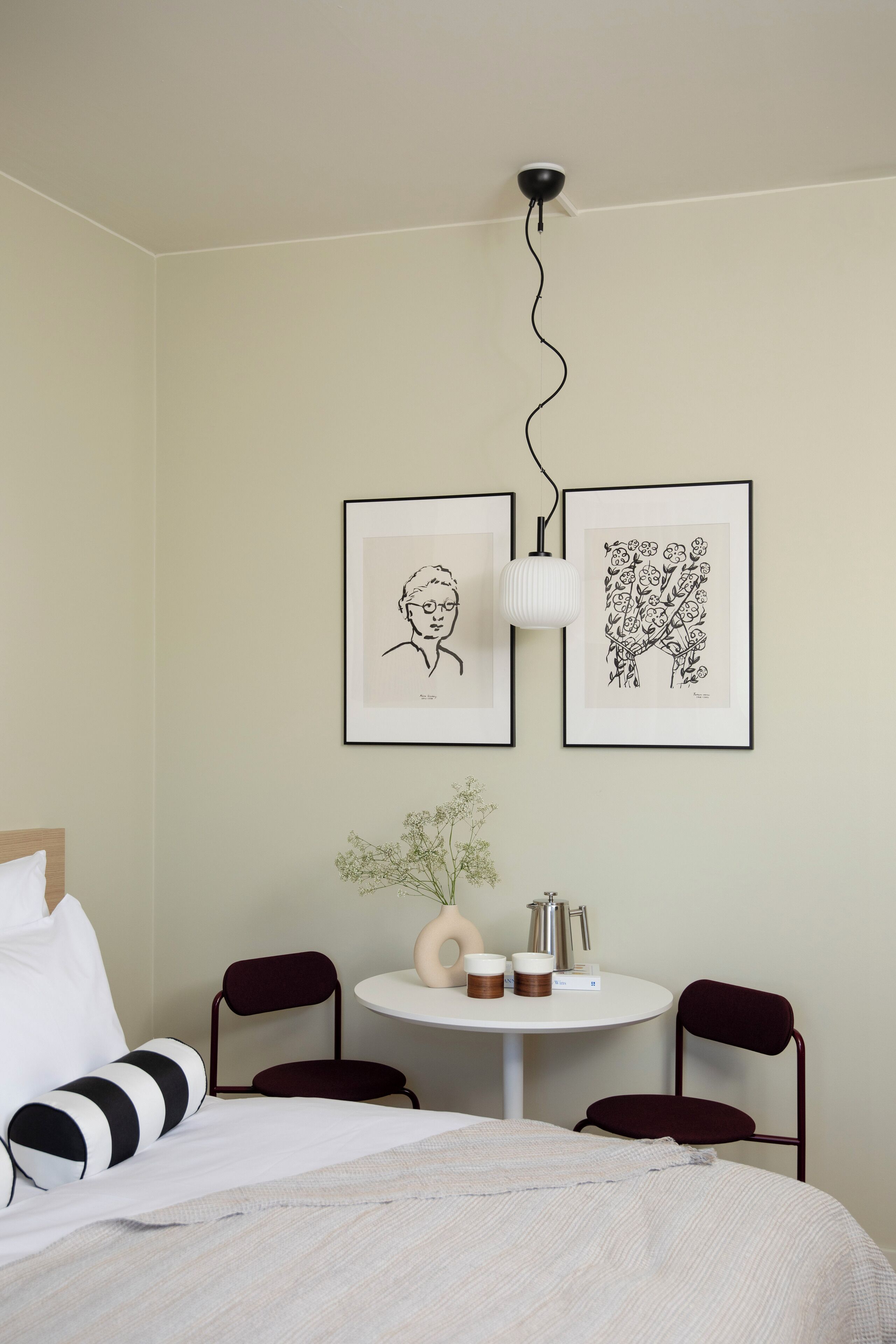 Et hotelværelse med en seng, to billeder i rammer, et lille rundt bord med en vase og to kopper, samt to stole.