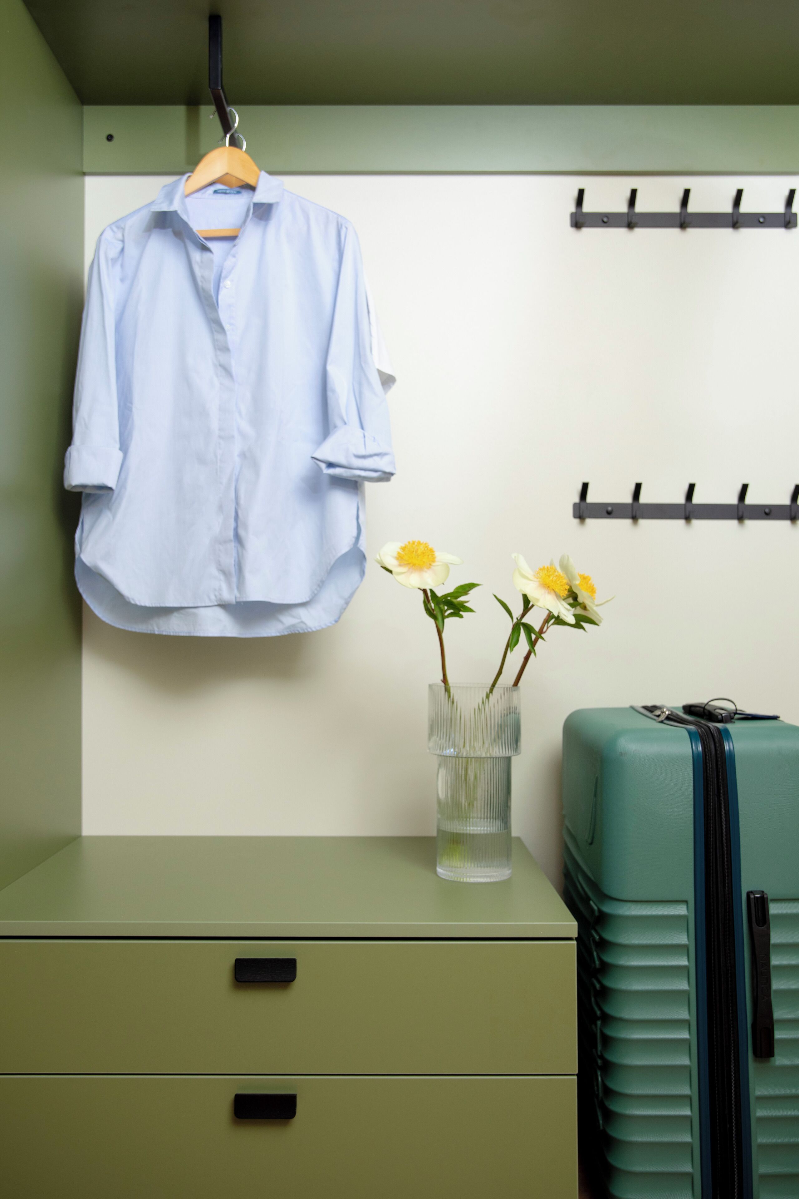 A room with a hanging shirt, a vase with flowers, a suitcase, and a filing cabinet.