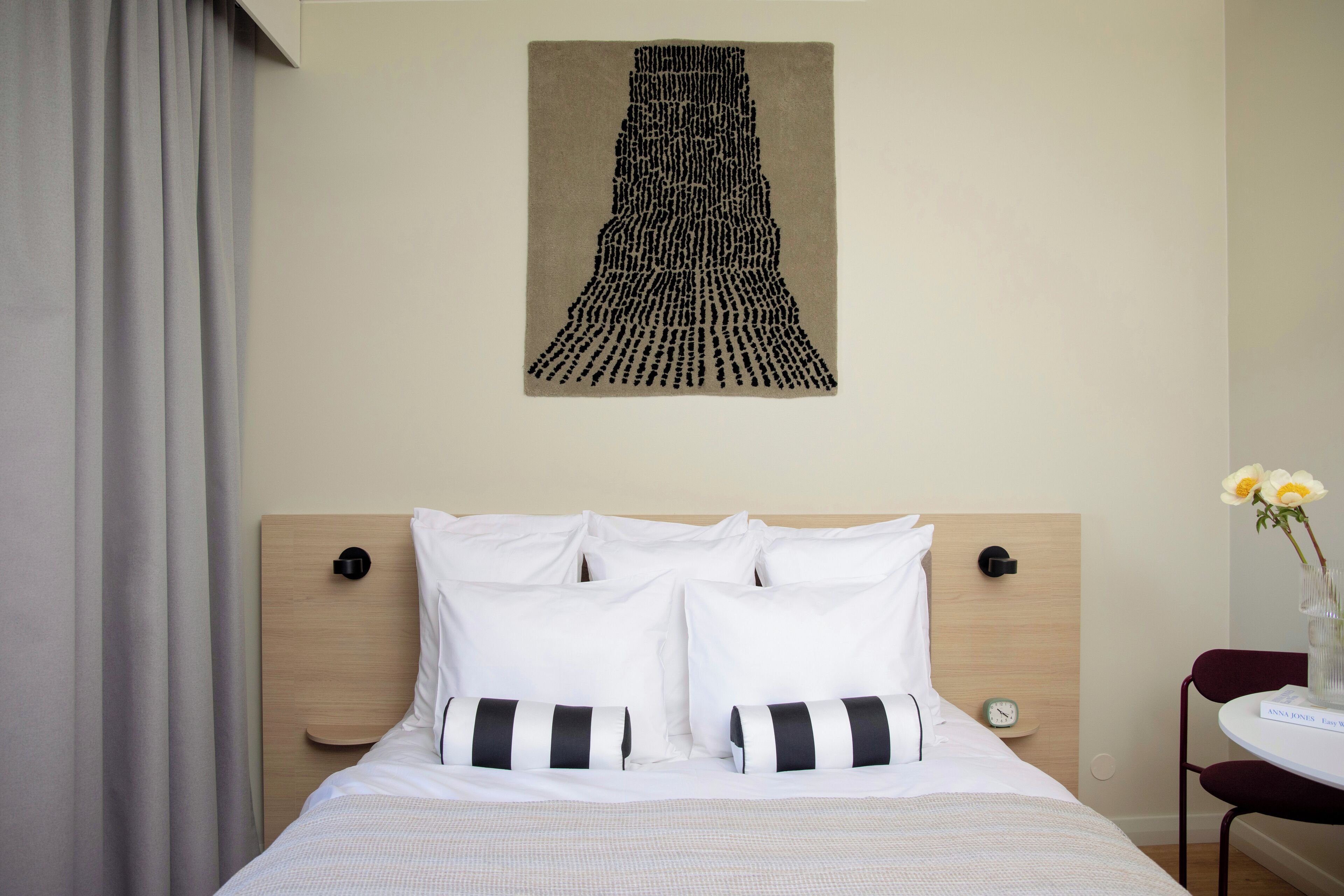 A hotel room with a bed, white bedding, a wooden headboard, a wall-mounted artwork, a bedside table with a vase of flowers, and a chair.