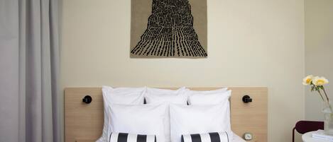 A hotel room with a bed, white bedding, a wooden headboard, a wall-mounted artwork, a bedside table with a vase of flowers, and a chair.