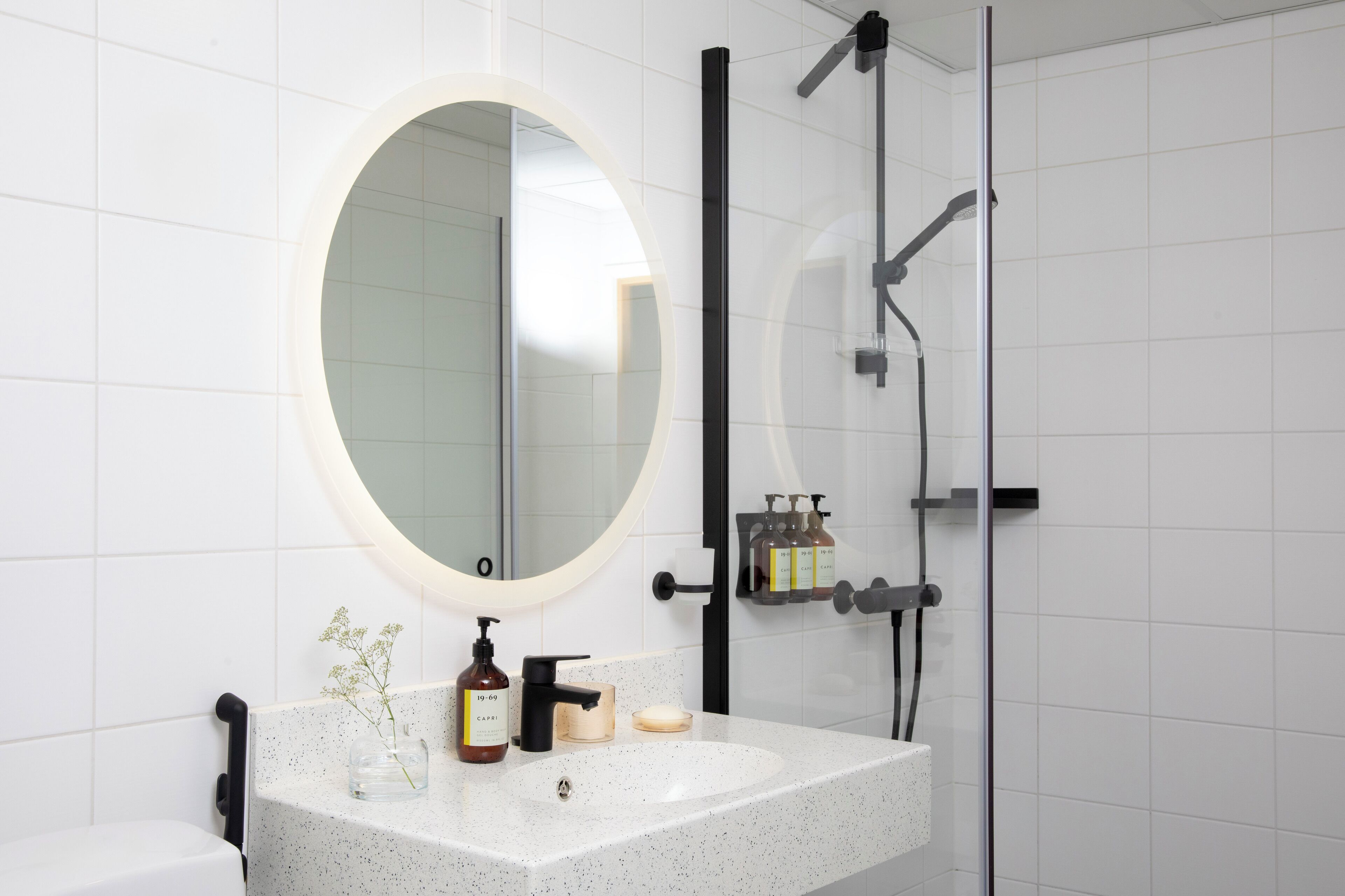A bathroom with a white tiled wall, a round mirror, a white sink with a speckled countertop, a handheld showerhead, and a glass shower enclosure.