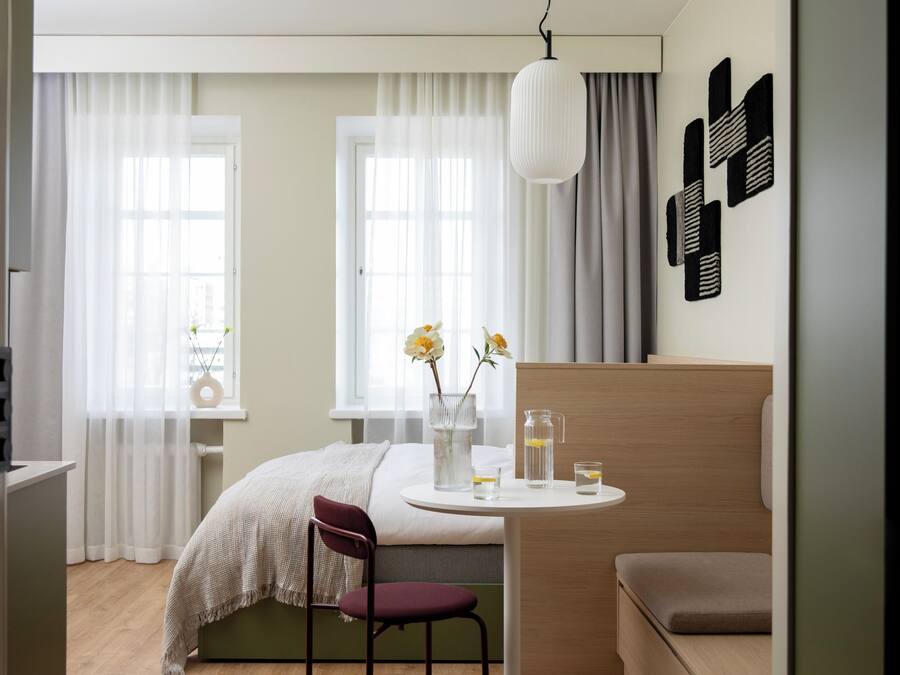 A modern bedroom with a bed, a desk, and a chair. There are curtains, a hanging light, and a wall decoration.