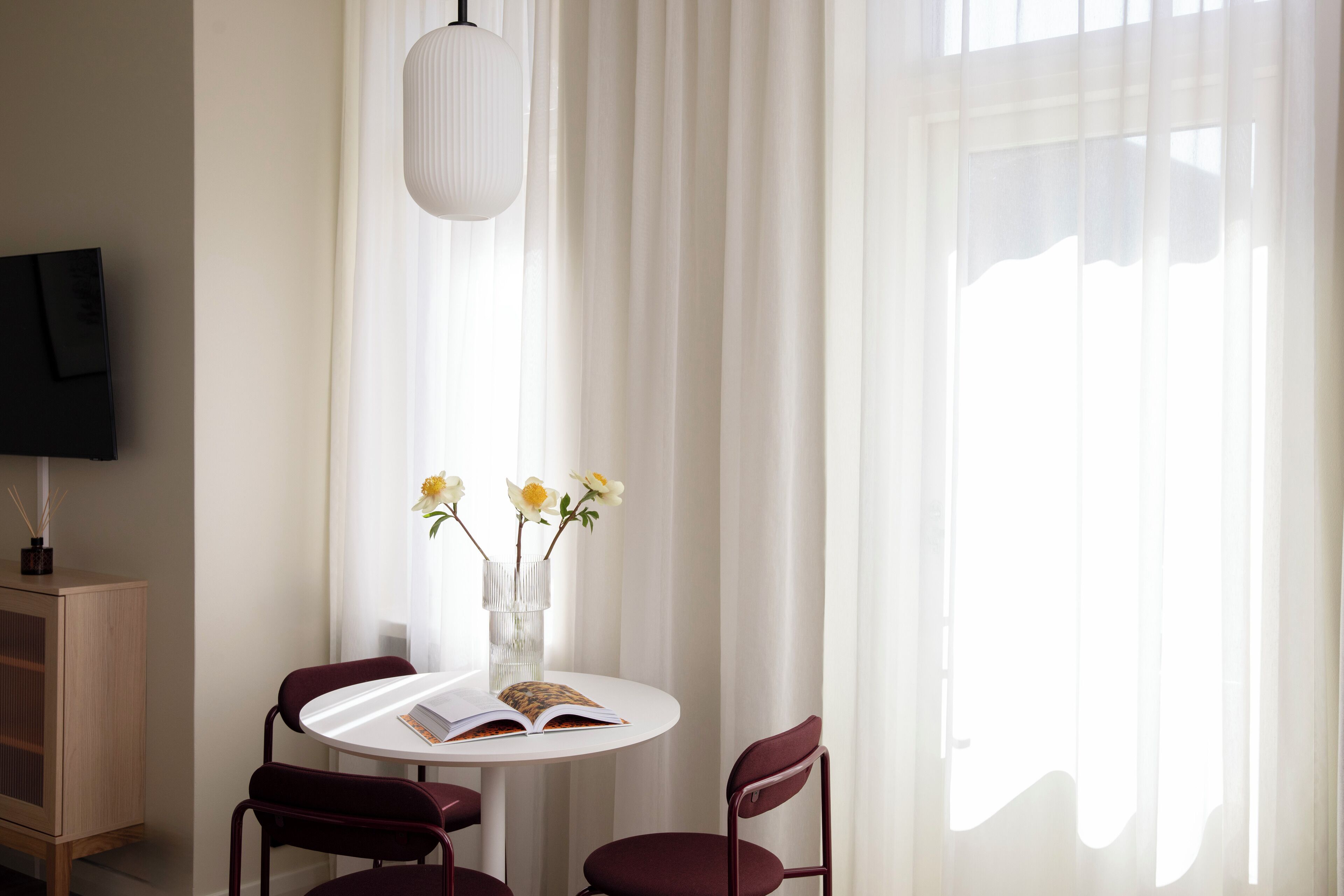 A modern room with a round table, two chairs, a vase with flowers, a book, and a television on the wall.