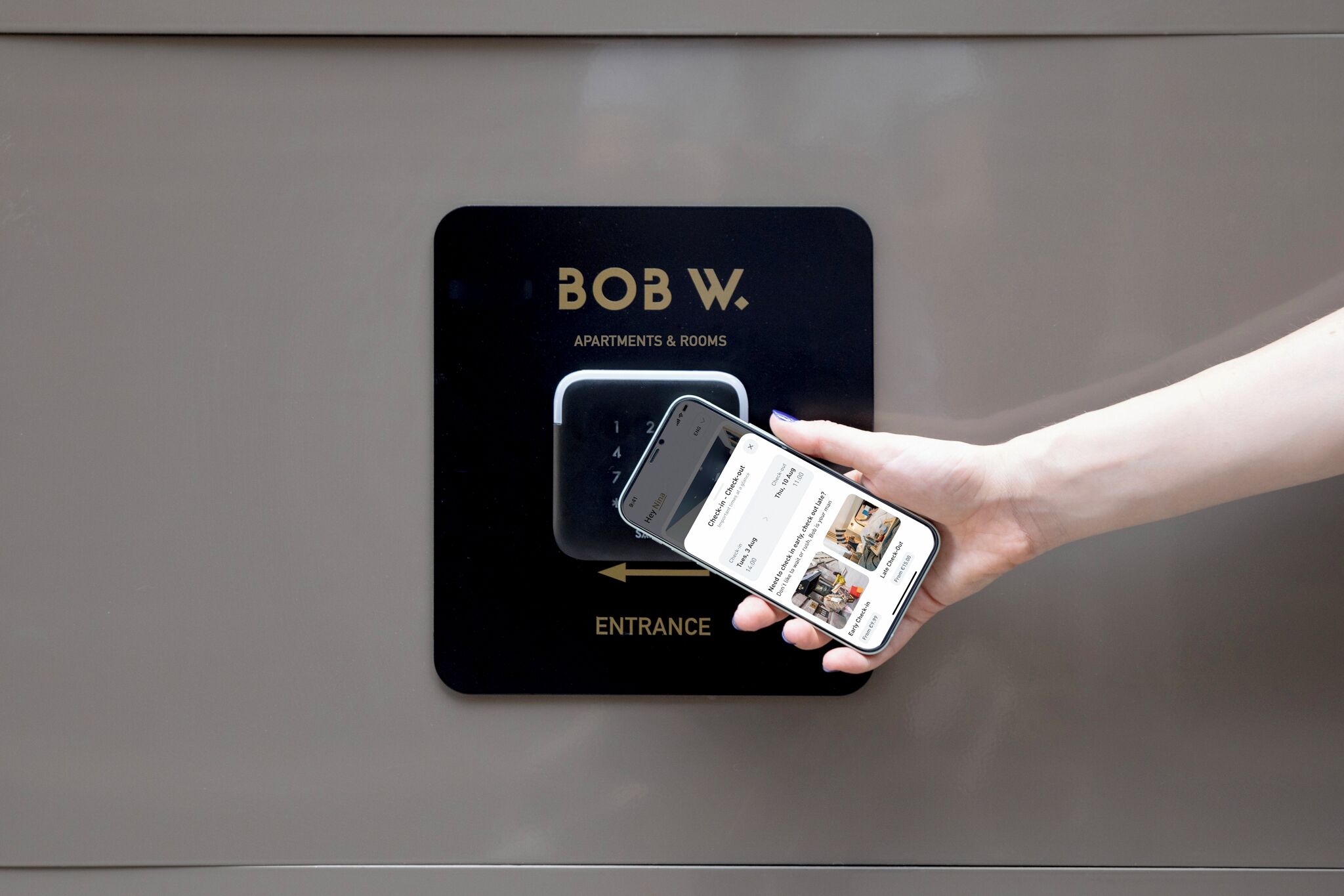 A hand holding a smartphone with a hotel app interface, pointing to a sign that reads "BOB W. APARTMENTS & ROOMS ENTRANCE."
