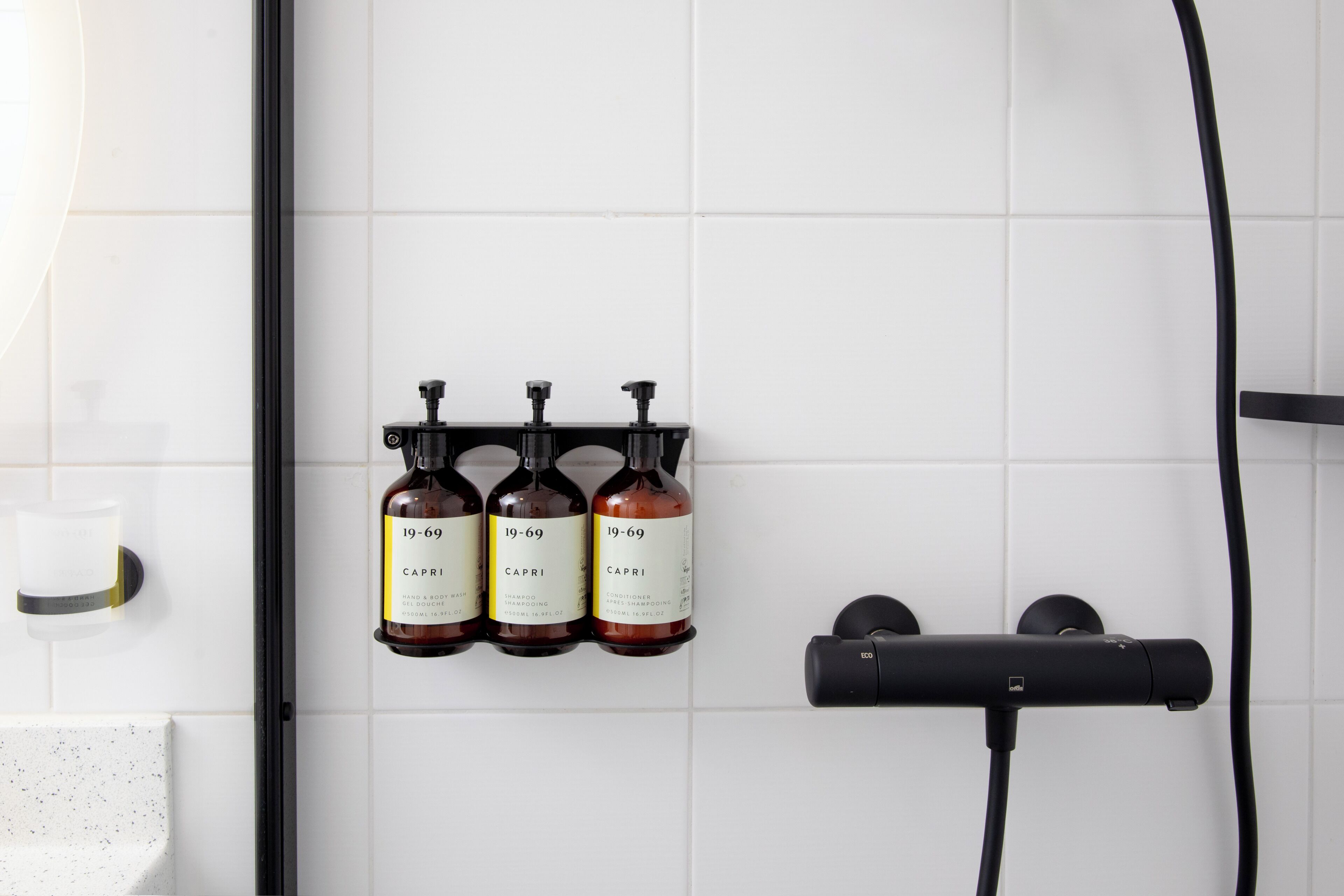 A bathroom with white tiles, three bottles of Capri shampoo and conditioner, and a black hairdryer on a black stand.