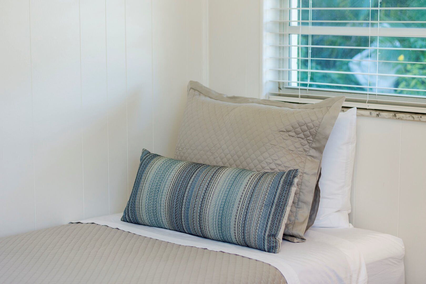 A neatly made bed with a striped pillow and a quilted pillow, set against a white wall with a window in the background.