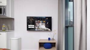 A modern hotel room with a flat-screen TV mounted on the wall, a wooden nightstand with a blue vase, and a window with curtains. - Bob W London Tower Hill  - Residences (London)