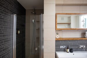Un baño moderno con ducha envuelta en vidrio, un lavabo blanco y un estante de madera con artículos de aseo.