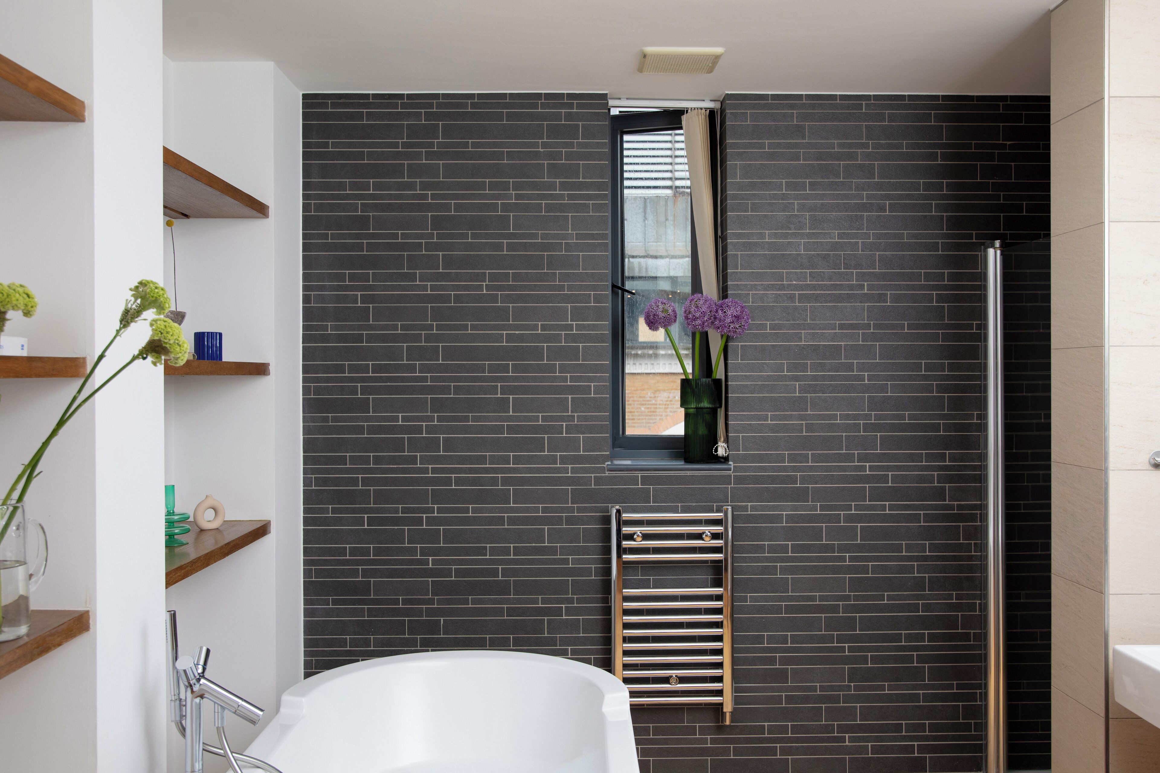 A modern bathroom with a black brick wall, a white bathtub, a glass shower enclosure, and a window with a view.
