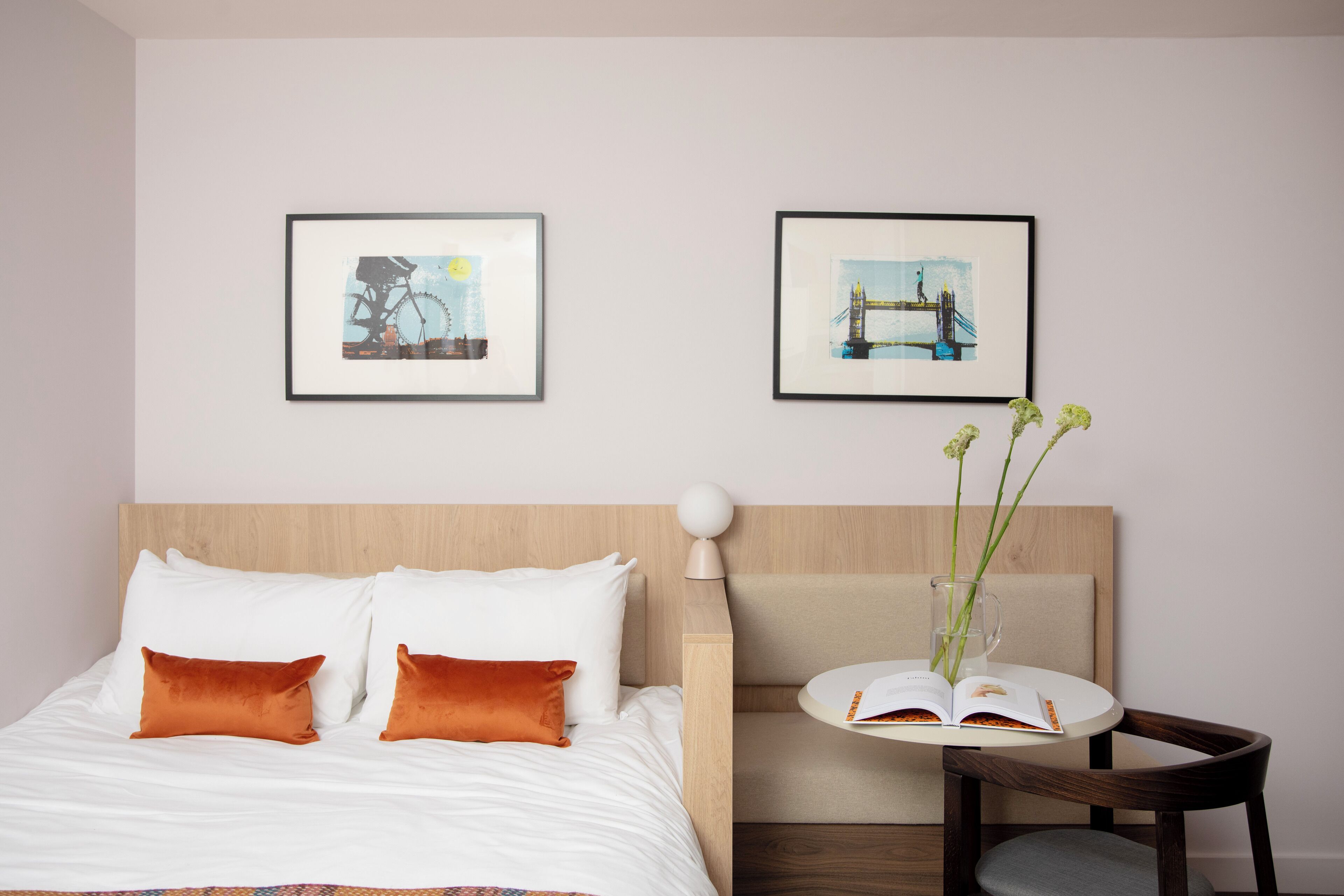 Ein Hotelzimmer mit einem Bett, zwei Kissen, einem Nachttisch mit Buch und Vase sowie zwei gerahmten Bildern an der Wand.