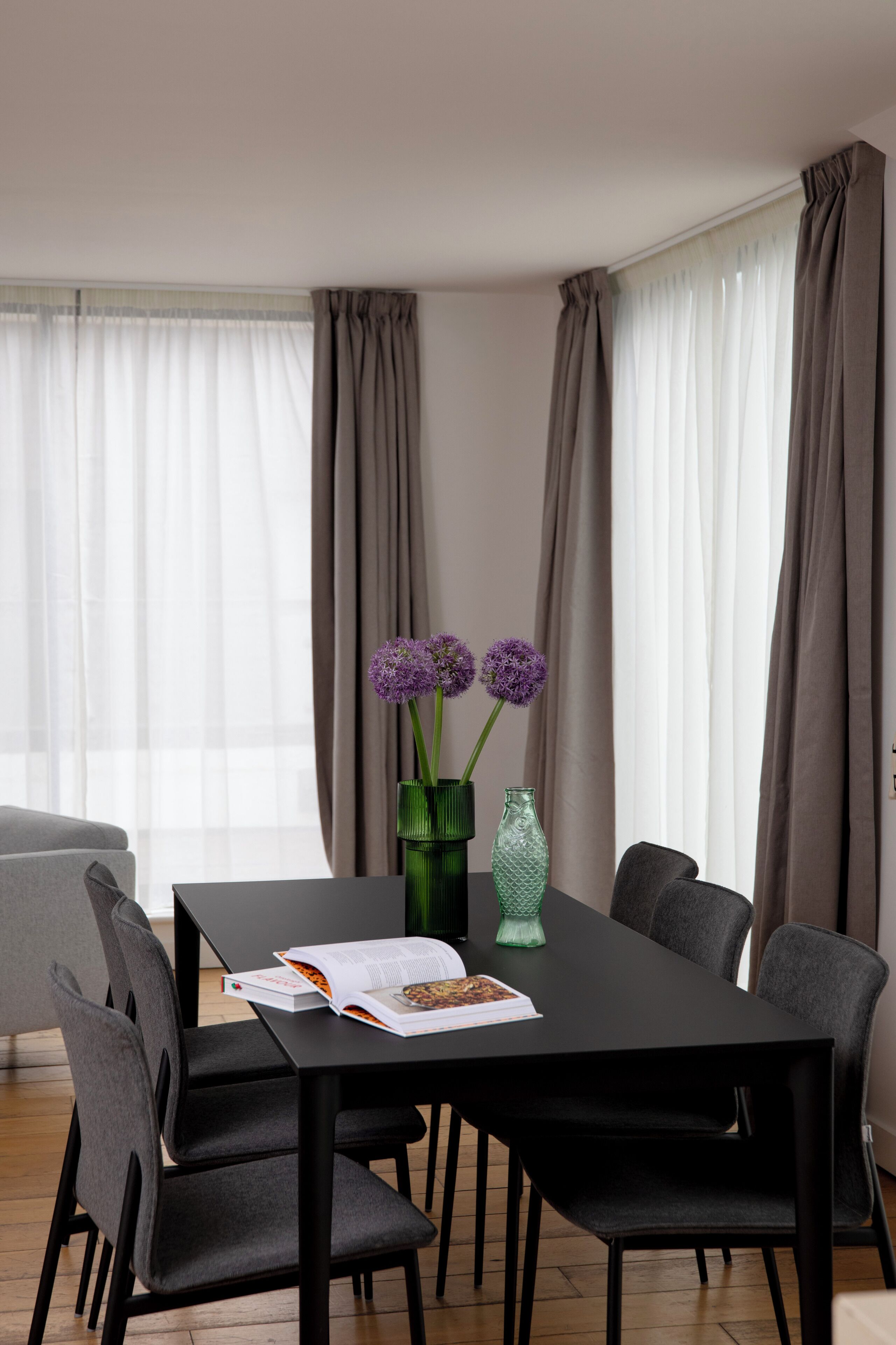 Ein moderner Essbereich mit einem schwarzen Tisch, sechs grauen Stühlen und einer Vase mit violetten Blumen.