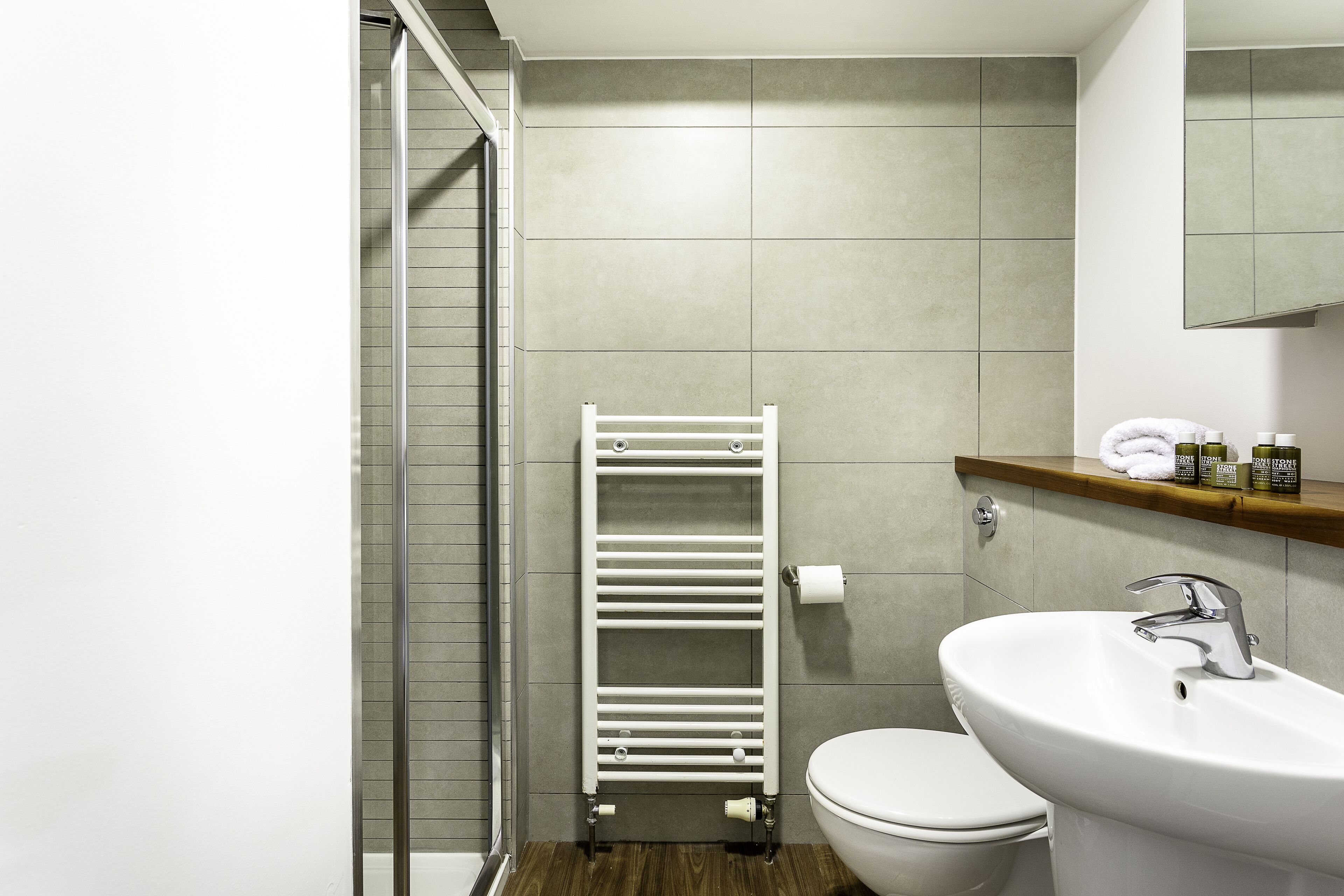 Un bagno con cabina doccia in vetro, un water bianco, un lavandino bianco con rubinetto cromato e uno scaffale in legno con articoli da toilette.
