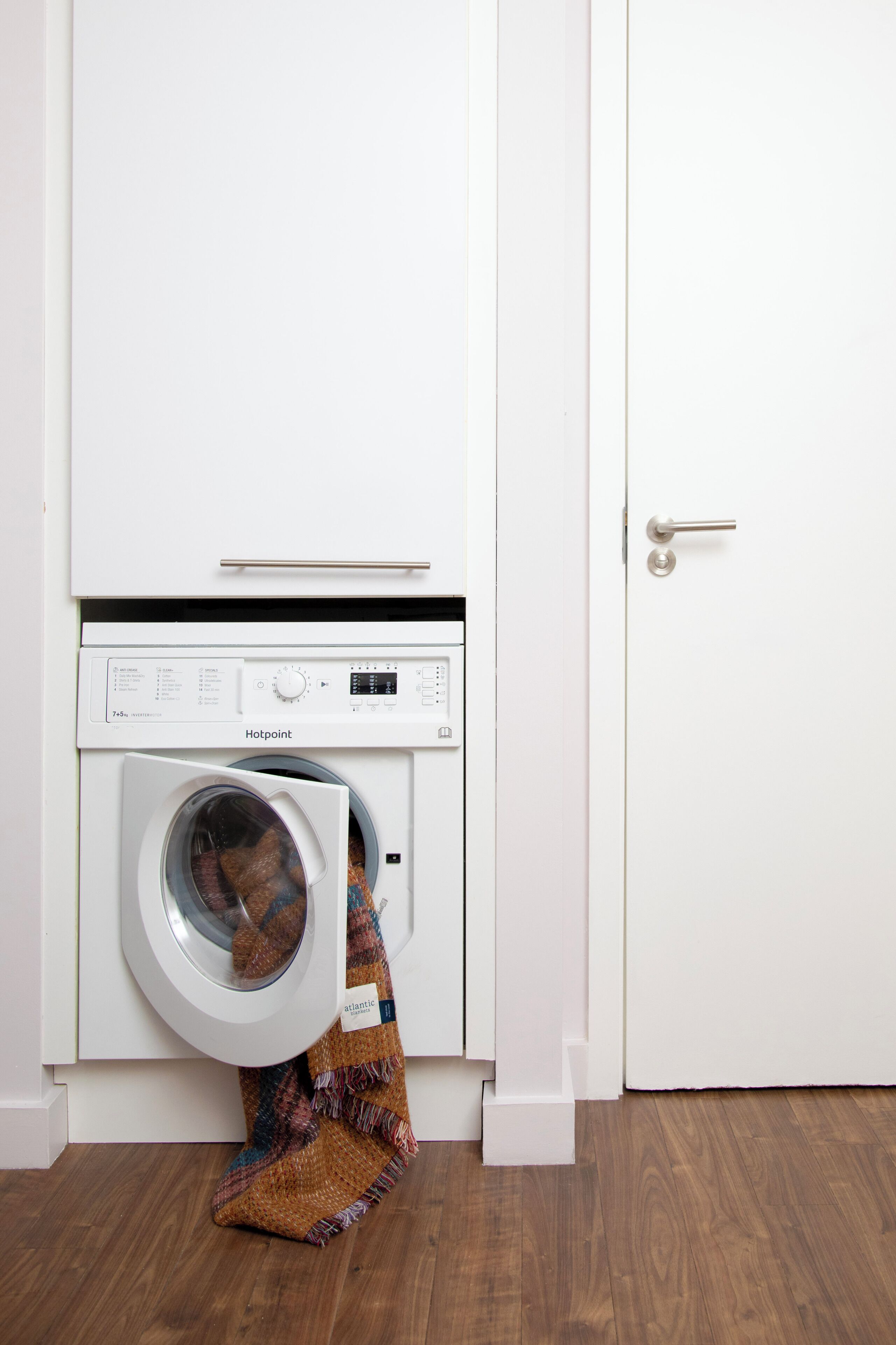 A white washing machine with a control panel, a patterned cloth hanging from the door, and a wooden floor.